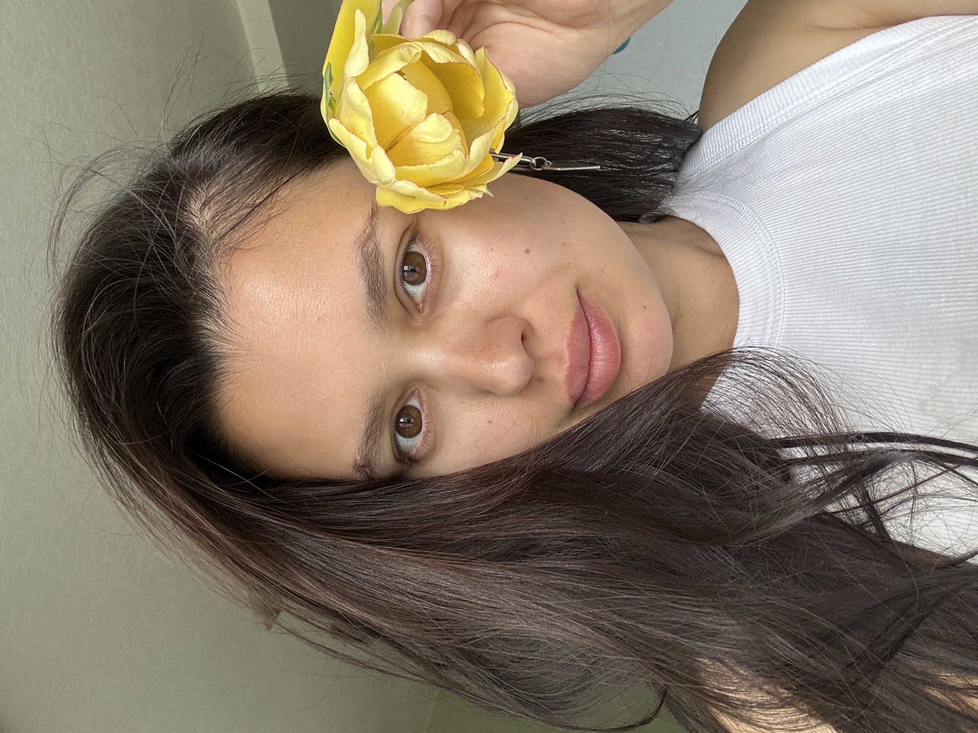 A woman with dark hair holds a yellow tulip near her face. She has brown eyes and fair skin, and is wearing a white shirt. Her dark hair is long and partly covers her forehead. The background appears to be a plain wall or surface, possibly indoors. There is no specific information to indicate this photo was taken in the city of Chaikovsky, Russia.