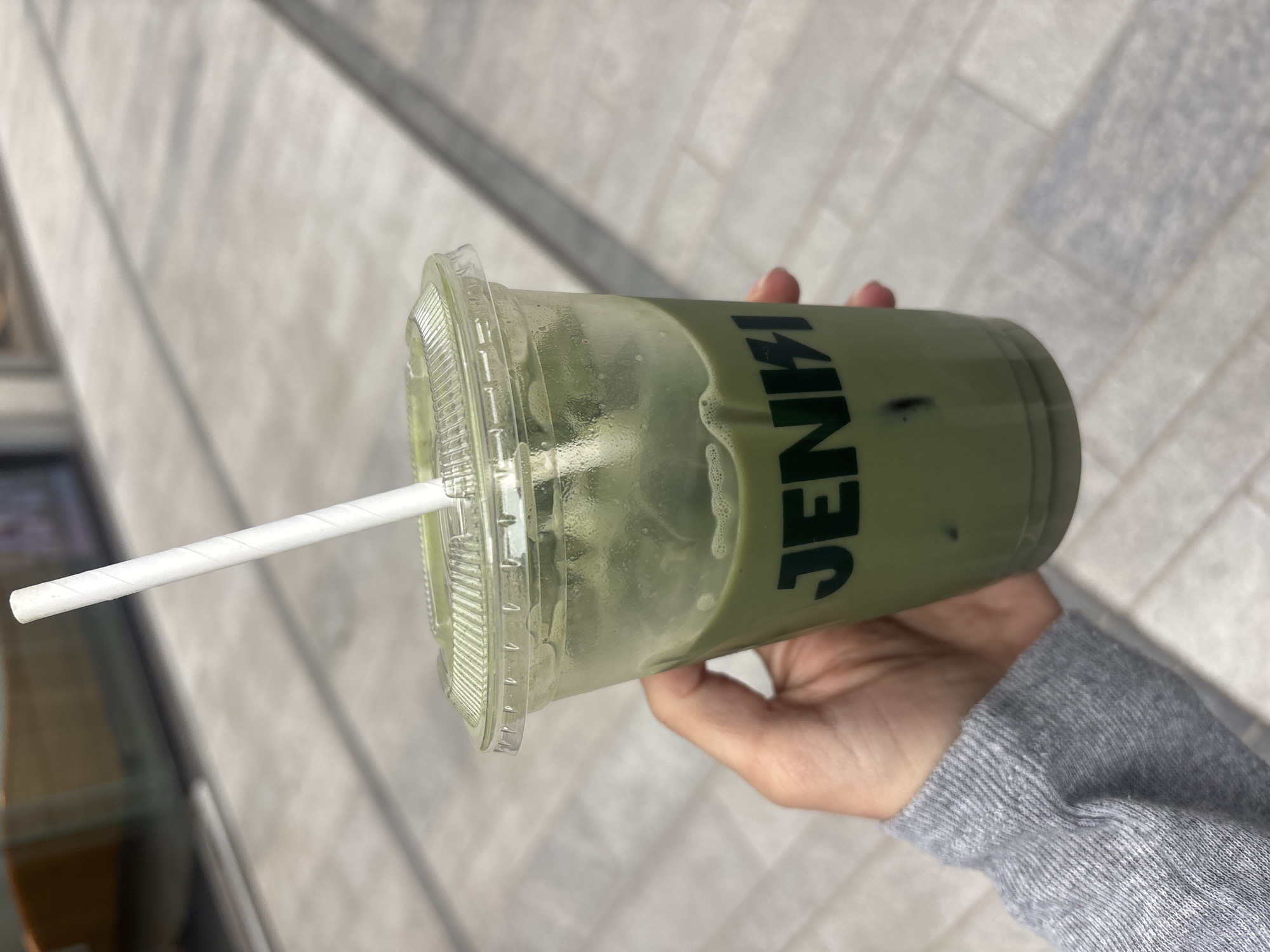A hand holds a Jenki matcha latte in a clear plastic cup with a lid and a white paper straw. The cup is condensation, indicating it is cold. The background shows paved surfaces suggesting an outdoor, urban setting. Given that Jenki is a London-based cafe, the location is likely within the city of London, United Kingdom. A grey-sleeved arm holds the drink.