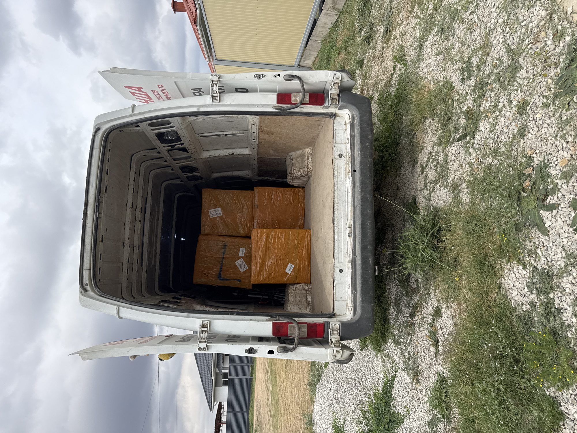 The image shows the rear view of an open white cargo van parked on a gravel and grass area, likely in the city of Uyutnoye, Ukraine. Inside the van, there are four large boxes wrapped in orange protective material, stacked two by two. The van doors are open, revealing the interior and the stacked cargo. In the background, a house with a dark roof and a grey fence is visible, along with a yellow corrugated metal fence. The sky is overcast.