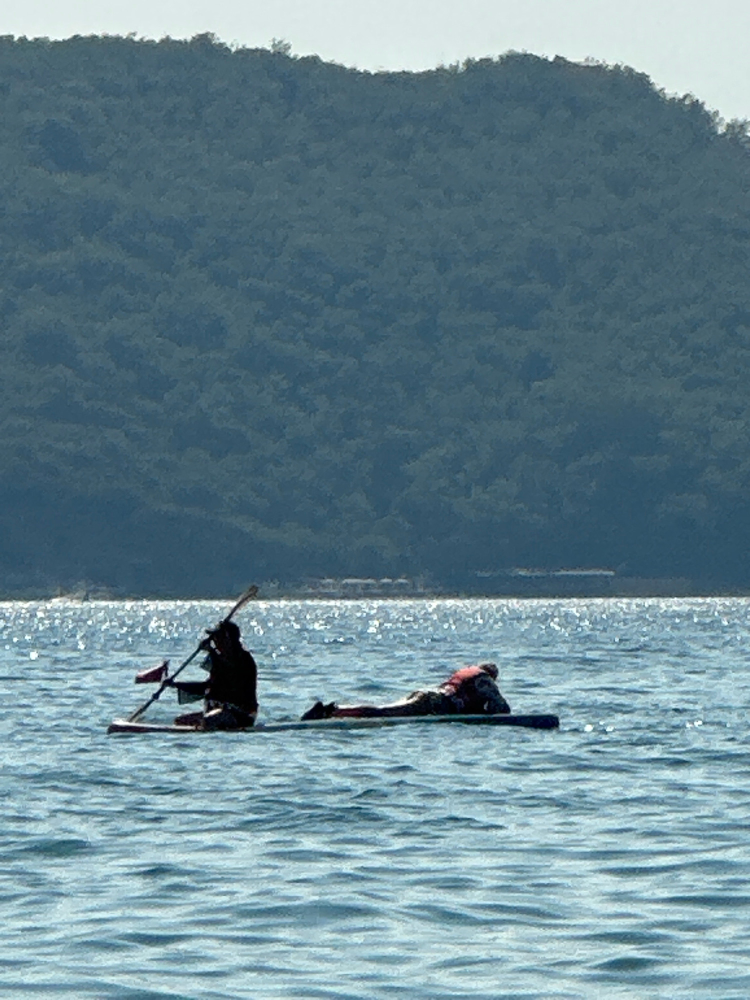 In Boreti, Montenegro, two individuals are engaged in water activities on a calm, sunlit sea. In the foreground, one person is seated on a paddleboard, holding a paddle. To their right, another person is lying prone on a separate paddleboard, wearing a life vest. The background features a lush, green, tree-covered hillside, typical of the coastal landscape of the region. The water reflects the bright sunlight, creating a shimmering effect.