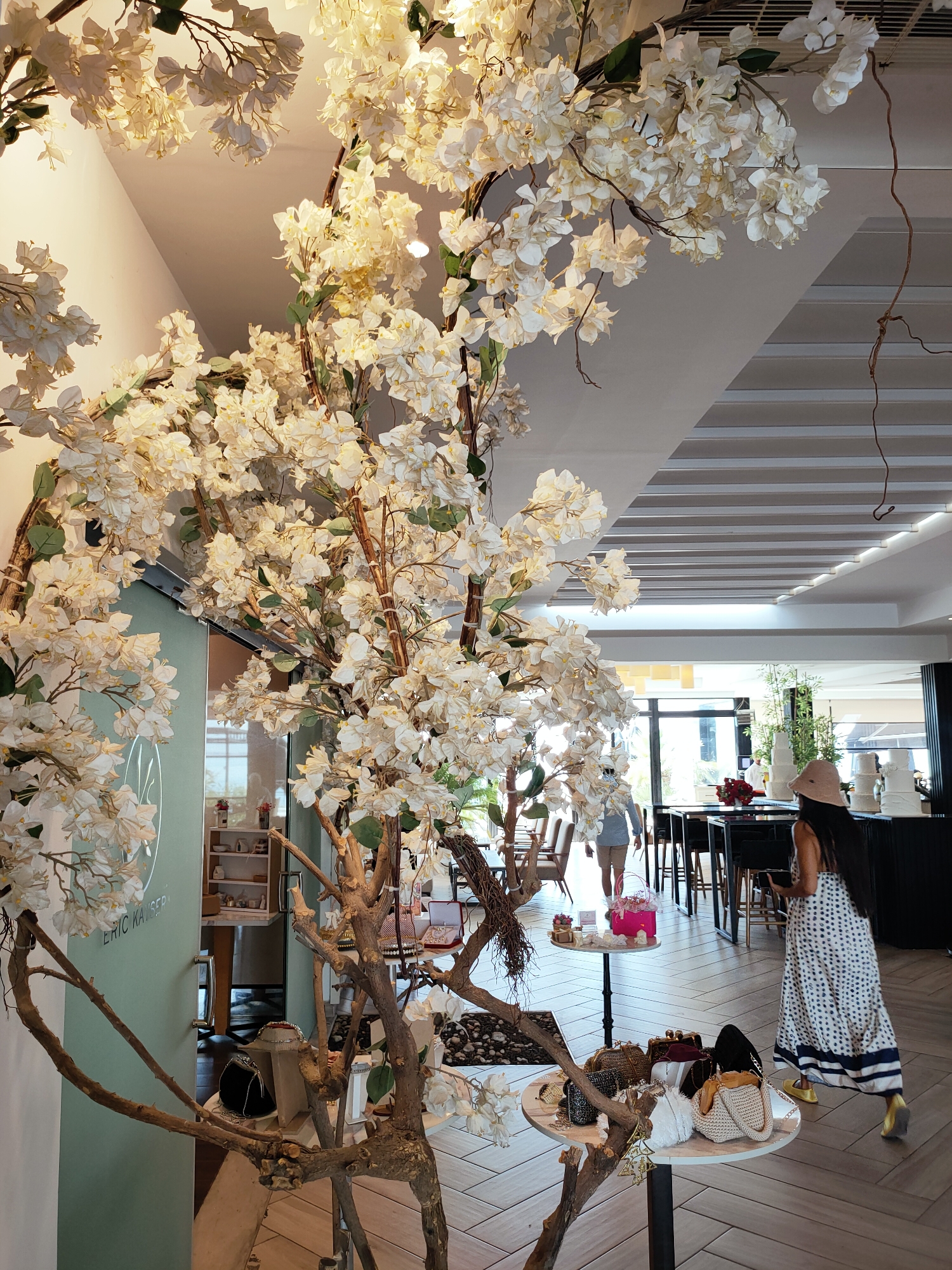 This is a full-shot, indoor photograph of a shop interior, potentially in Tunis, Tunisia, characterized by an elegant and decorative ambiance. Dominating the foreground and midground are large, artificial white flowering branches, possibly bougainvillea, which are artfully arranged to create a dramatic and visually appealing display. These branches extend from the bottom left and upper portions of the frame, with delicate white flowers and some green leaves.
In the background, the scene unfolds within a well-lit and modern retail space. To the left, a teal-colored wall features some text, partially obscured by the floral arrangement, suggesting a brand name or store title. A glass door or partition is visible, offering a glimpse into an adjacent area with shelves displaying small items.
Further into the background, a woman with long dark hair, wearing a light-colored wide-brimmed hat and a long, patterned dress with blue and white polka dots, walks away from the camera. She is wearing yellow shoes and appears to be browsing or moving through the store. To her right, on a counter or display area, several tiered white cakes are visible, suggesting a bakery or patisserie section. Behind the cakes, there are tall green plants, adding a touch of natural greenery to the setting.
In the mid-right of the image, several small round tables are arranged. One table closer to the viewer displays a collection of handbags and clutches in various styles, textures, and colors. Another table further back has a pink box with a bow and some smaller items, possibly gifts or accessories. The flooring consists of light-colored rectangular tiles arranged in a herringbone pattern. The ceiling is modern, with recessed lighting and a series of parallel beams or panels. The overall impression is one of a stylish and tastefully decorated establishment.