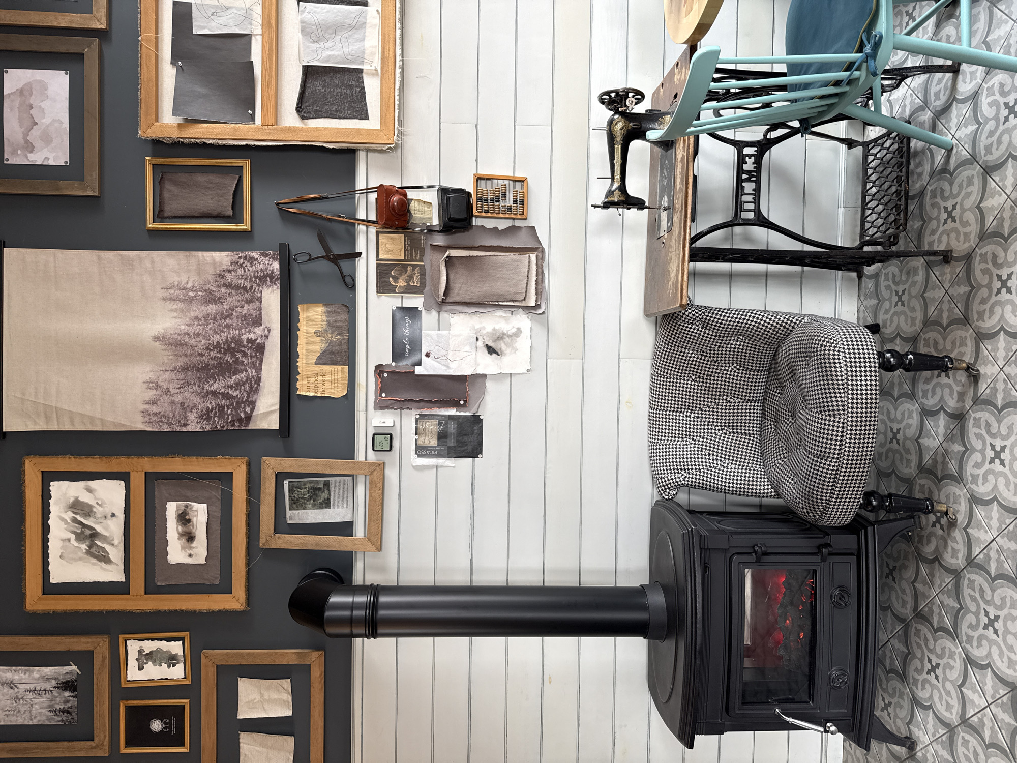 This overhead shot captures an interior scene, likely a creative studio or cozy living space in Krasnoyarsk, Russia. The room is divided visually by different wall treatments and floor coverings.
On the left, a dark gray wall is adorned with numerous framed items. These include abstract watercolor paintings, a large framed textile print depicting a snowy forest, and smaller framed fabric swatches and newspaper clippings.
A central white planked wall serves as a backdrop for a curated collection of objects and a mood board. Here, various fabric swatches in earthy tones, sketches of figures, small photographs, and torn paper pieces are pinned. Visible text includes "simple therapy" on a white card, "PICASSO" on a black card (partially obscured), and fragments like "...an art..." and "...the world?" on another. Vintage items are also displayed: a pair of scissors, a brown leather-cased camera, a black box camera, and a wooden abacus. A small digital display on the wall shows "22°".
The floor in the foreground features gray and white patterned tiles, while a section near the white wall has white plank flooring. A prominent black wood-burning stove, with a visible glowing fire inside, provides a focal point. Adjacent to the stove is an armchair upholstered in a black and white houndstooth pattern. Further to the right, a vintage sewing machine table with a cast-iron base, marked with the Cyrillic text "ПОДОЛЬСКЪ" (Podolsk), is positioned with a light blue wooden chair. The scene evokes an atmosphere of warmth, creativity, and vintage charm.