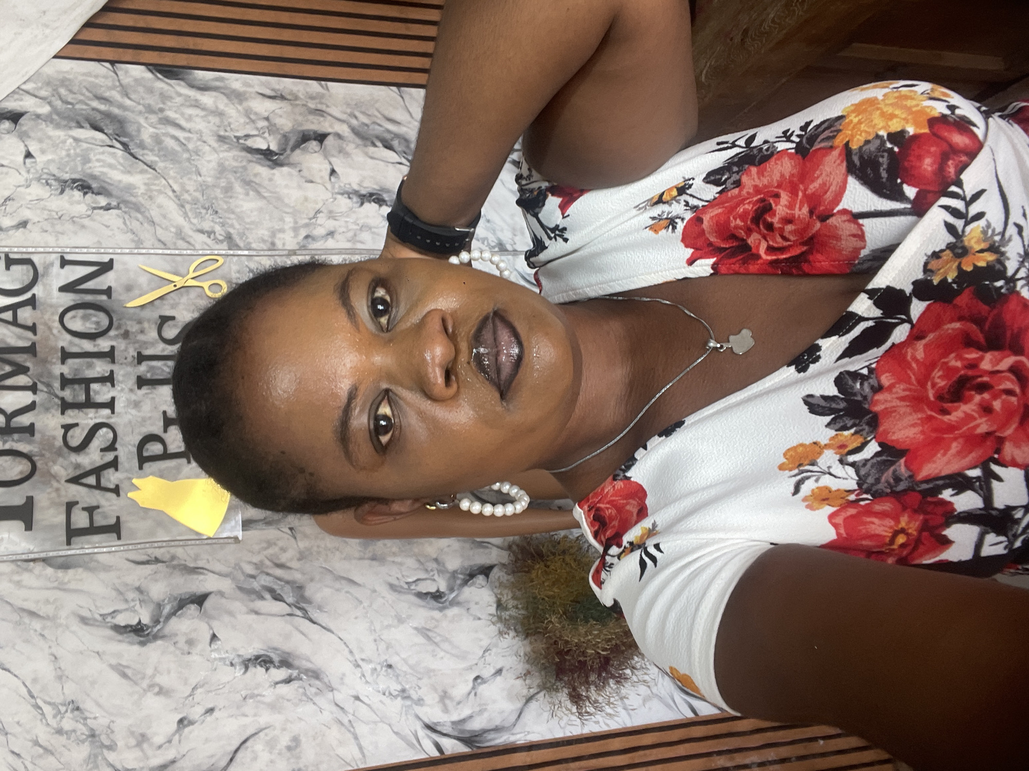 A medium shot shows a woman with dark skin and dark hair, pulled back from her face. She is wearing a white dress with a floral pattern of red, yellow, and black flowers. She has large pearl hoop earrings and a silver necklace with a small apple-shaped pendant. Her lips are painted with dark lipstick. She is looking at the camera with a neutral expression. Her right arm is behind her head. The background consists of a wall with a marble pattern and vertical wooden blinds. A sign with the words "TORMAG FASHION PLUS" and graphics of a yellow dress and gold scissors is visible behind her. A dried plant arrangement is to her left. The image is a selfie taken from a slightly elevated angle. The scene is likely indoors, possibly in a salon or shop, given the signage. Badagry, Nigeria, is mentioned as the location.