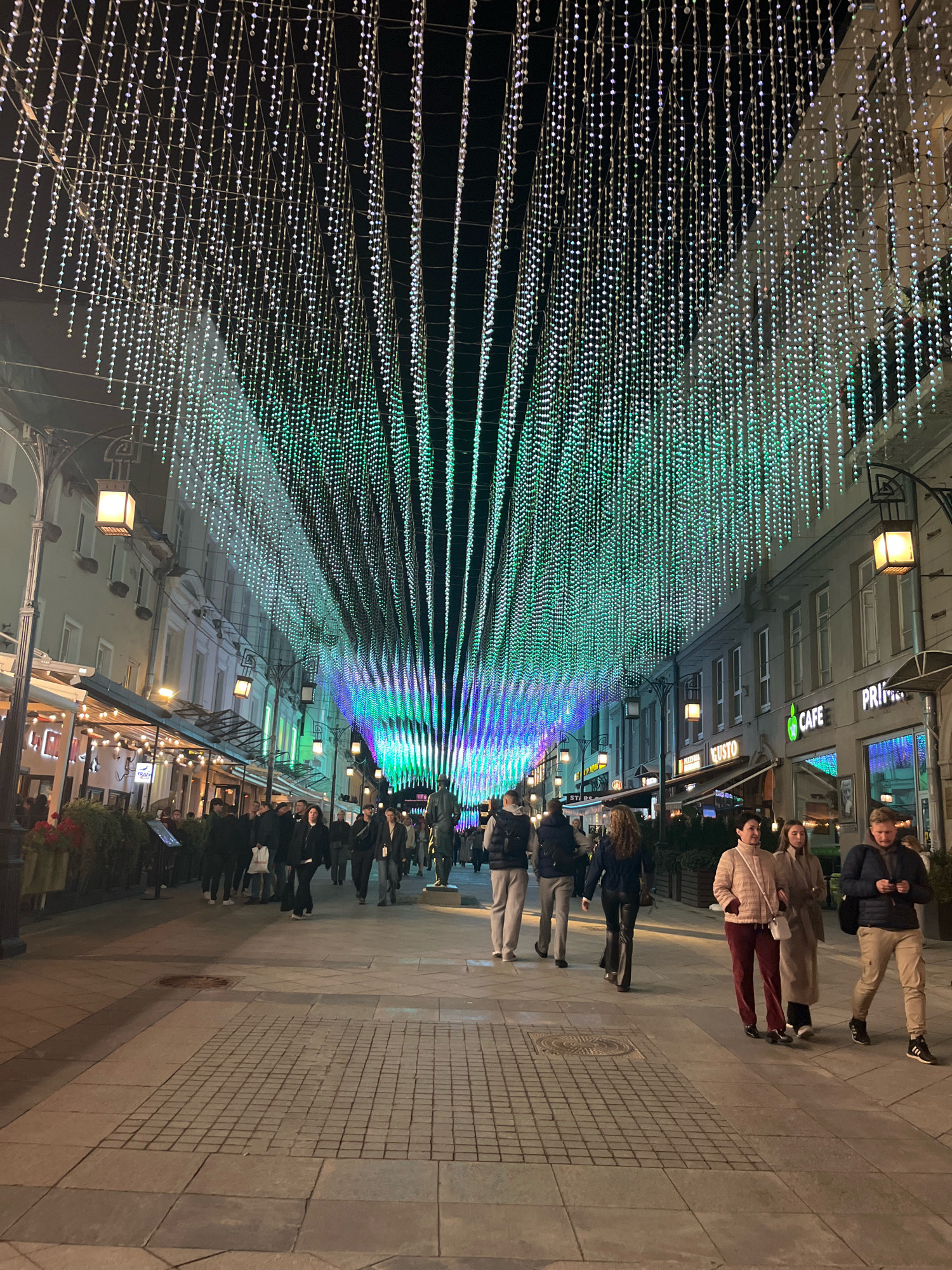 The image depicts a bustling pedestrian street in Moscow, Russia, at night, adorned with a spectacular display of illuminated string lights. The lights, forming a canopy over the street, transition in color from white and blue to green and purple, creating a vibrant, tunnel-like effect. Along the sides of the street, buildings with illuminated windows and storefronts line the way, suggesting a commercial or entertainment district. Several people are visible walking along the street, some in groups, others individually, dressed in casual winter attire. Shops and cafes with outdoor seating are present on the left side of the street, with patrons seated at tables. A statue of a man stands to the right, adding a cultural element to the urban landscape. The street is paved with cobblestones, and lampposts with square lanterns are strategically placed along the pathway. The overall atmosphere is one of urban vibrancy and festive illumination, characteristic of a popular city destination during an evening.