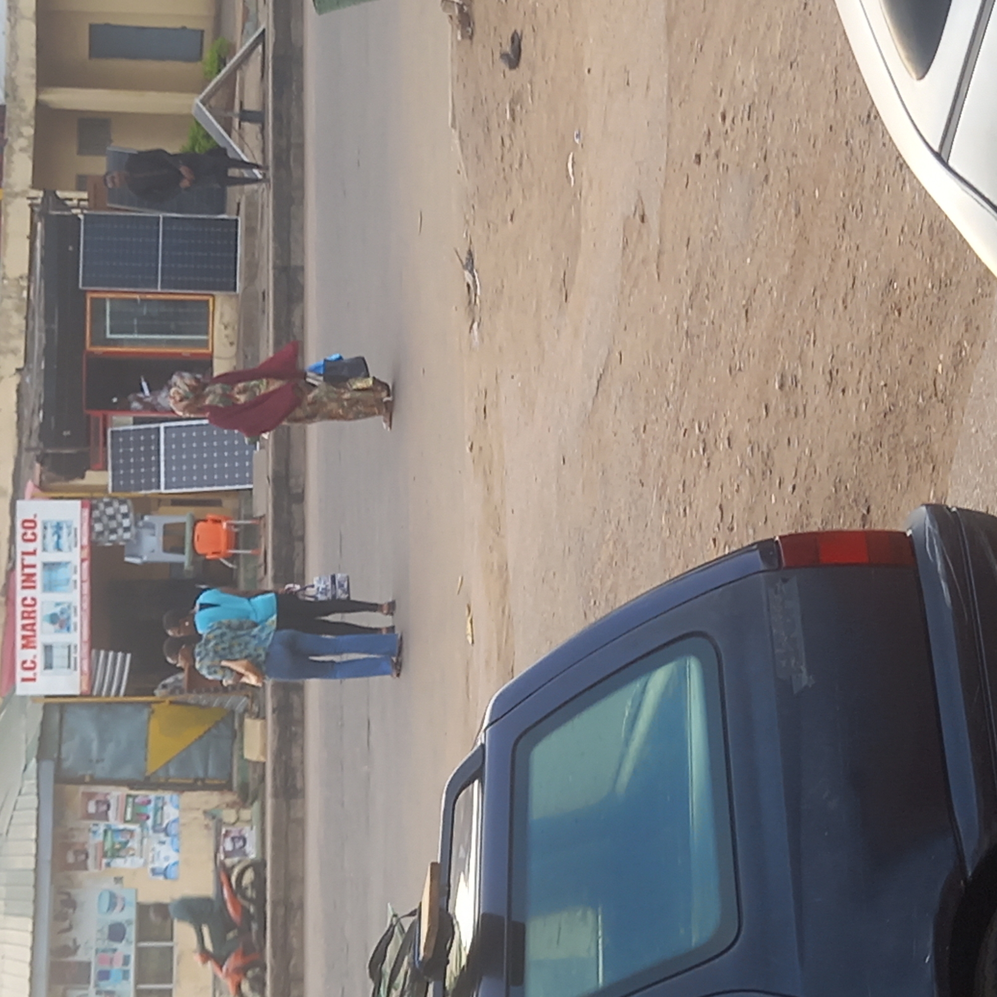 The image depicts a street scene in Dadi Riba, Nigeria. Several people are present, including women walking on the side of the road. A dark blue car is prominently featured in the foreground, with a white car visible partially in the upper right corner. A storefront for "I.C. MARC INT'L CO." is visible on the left. The street is unpaved. Solar panels are on display outside of the I.C. MARC INT'L CO. storefront.