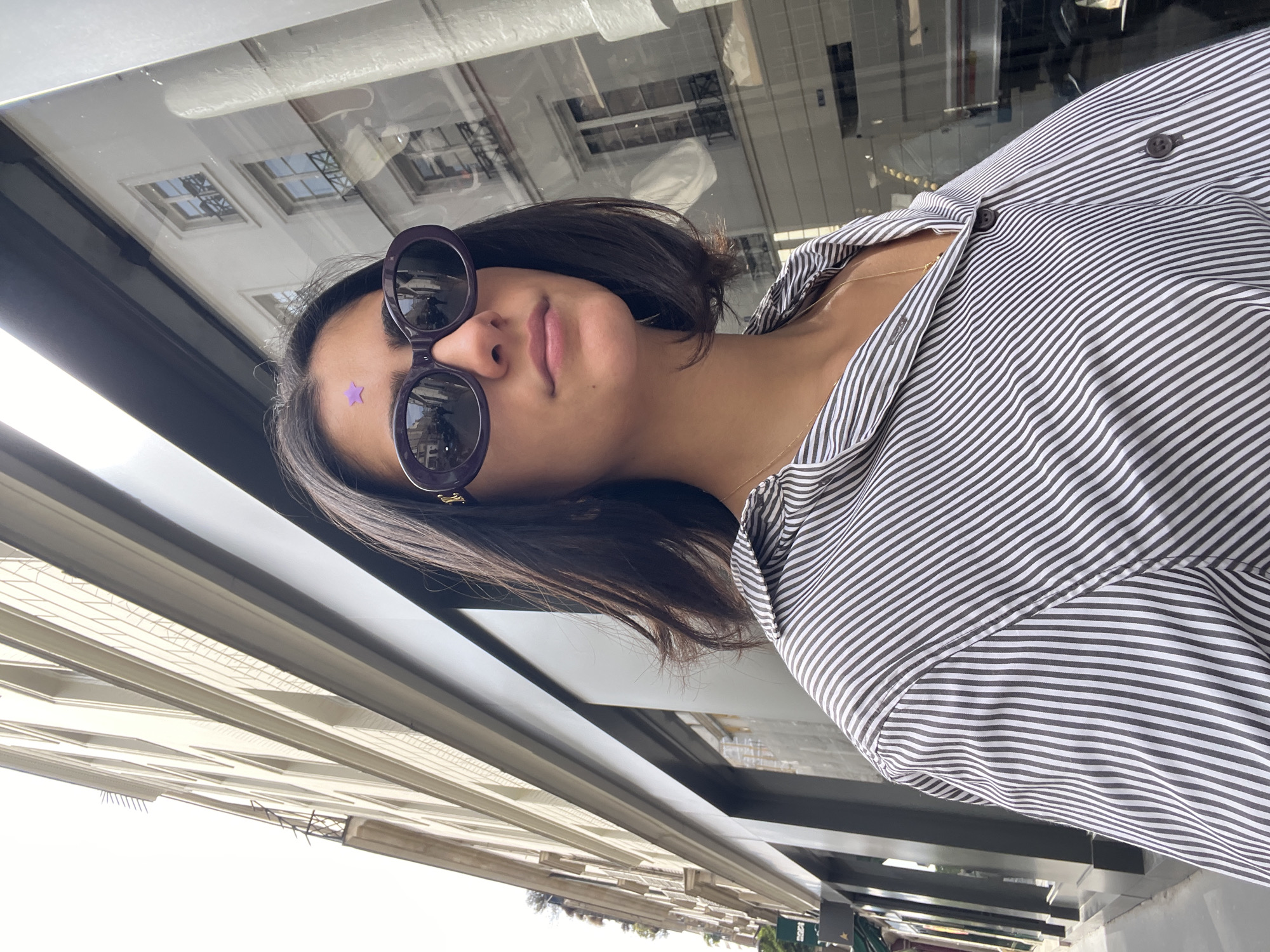 A woman with sunglasses is standing outside in Paris, France. The photo appears to have been taken through a window or from a raised perspective. The woman has short, dark hair and is wearing a striped shirt. She has a small purple star sticker on her forehead. The sunglasses have dark lenses and a purple frame. Reflections of buildings and windows can be seen in the sunglasses. The background shows Parisian architecture, with buildings featuring windows and balconies. Below the woman, there's a view of a street with greenery and a few signs or structures. The light indicates it is daytime.