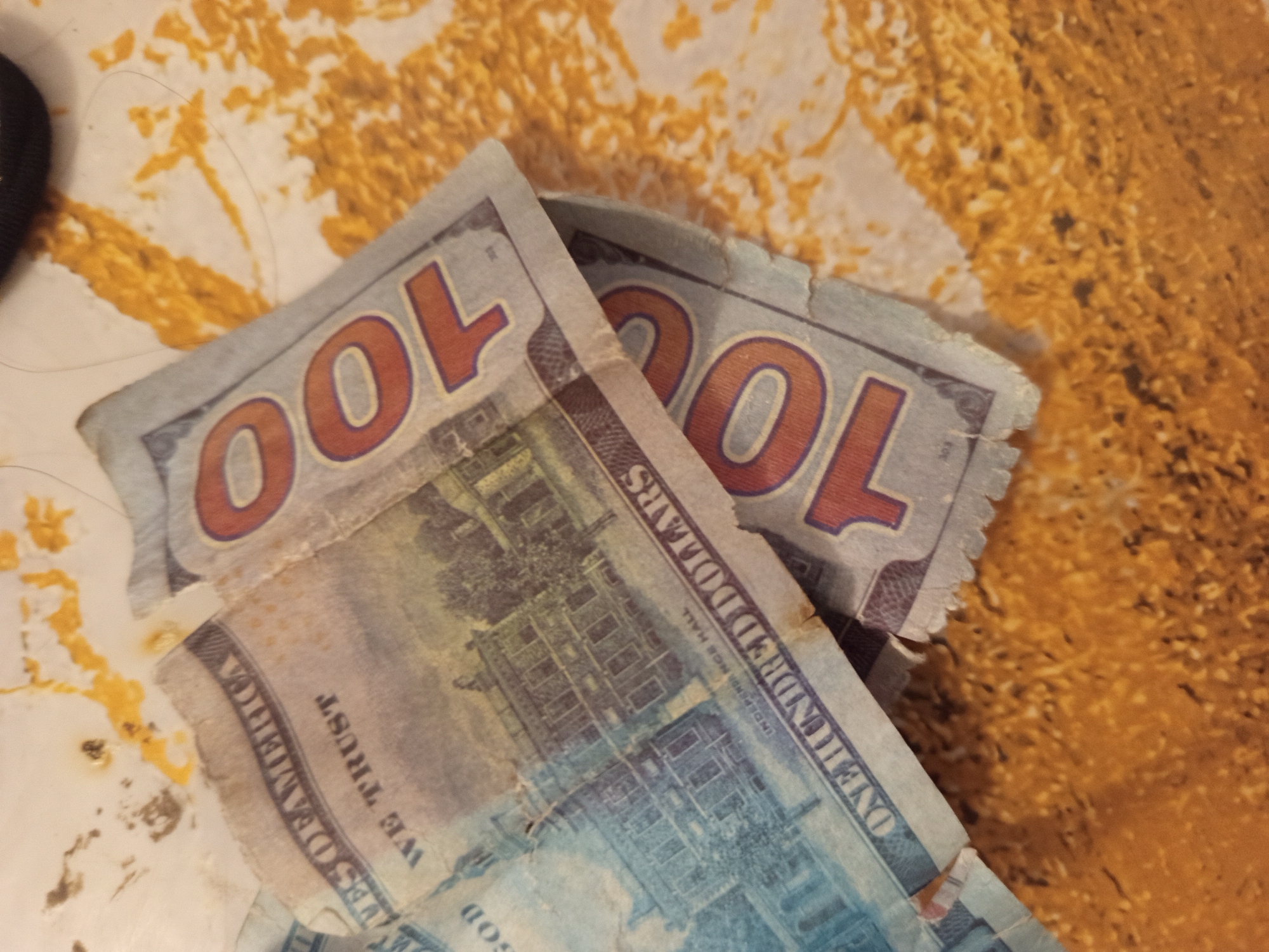 The image displays two US hundred-dollar bills, partially crumpled and placed on a textured surface with a marbled or speckled pattern of yellow and white. The bills are oriented diagonally, with the denomination "100" prominently visible in orange on the bottom bill. The text "ONE HUNDRED DOLLARS" is partially visible on both bills. The background surface appears to be a table or countertop. There are no people or signs of activity visible in the image, and the scene does not provide any information that would allow for localization to Jardy-Su, Kyrgyzstan.