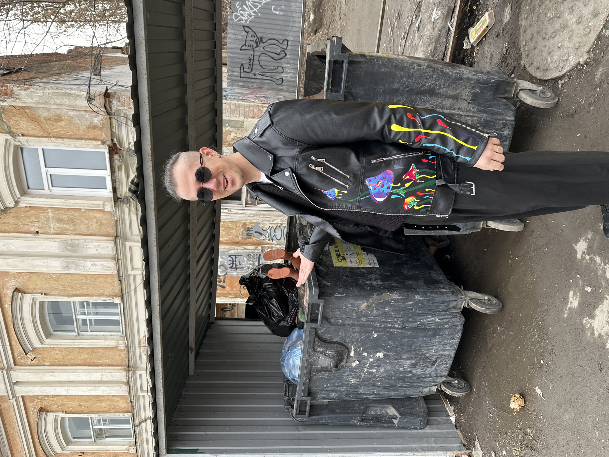 The image captures an adult male posing in an urban outdoor setting in Saratov, Russia. The man stands slightly left of center, facing the camera with a smile. He has short, light-colored hair with shaved sides, wears dark round sunglasses, a black leather jacket adorned with colorful mushroom and drip designs, a black sweater, and black pants. A silver ring is visible on his right ring finger. His right hand is extended, palm open, towards a large, dark-colored wheeled trash bin, as if presenting its contents.
The trash bin is located within a metal-framed enclosure with a corrugated metal roof. The bin is full of refuse, including black and blue plastic bags, from which two bare human-like feet, appearing to belong to a doll or mannequin, protrude vertically. Another similar trash bin is partially visible to the right. The ground in the foreground is muddy, uneven, and covered with dirt and scattered debris, including a crumpled piece of brown paper. A metal manhole cover is partially visible on the right.
In the background, a multi-story building with a faded orange stucco facade, white decorative cornices, and arched window frames shows signs of disrepair with peeling paint and cracks. Modern white-framed windows are installed in the building. To the right of this building, a corrugated metal fence or wall is heavily covered in graffiti, including a stylized drawing of a four-legged animal. Bare tree branches are visible against the sky in the upper right background.
