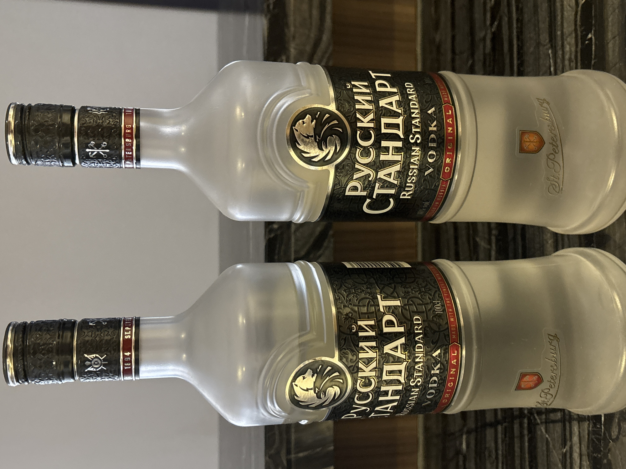 Two bottles of Russian Standard Vodka stand side by side on a dark, textured countertop. The bottles are made of frosted glass and have a distinctive shape, tapering inwards towards the neck and then flaring out slightly at the base. Both bottles feature the same label, which includes the Russian Standard logo, Russian text that translates to "Russian Standard," and English text that reads "Russian Standard Vodka." The labels also indicate "100cl" and "Original." The lower portion of the bottles is imprinted with the words "St. Petersburg" and a crest. The background consists of a plain, light-colored wall. There is no indication that this scene is located in Dubai, United Arab Emirates.
