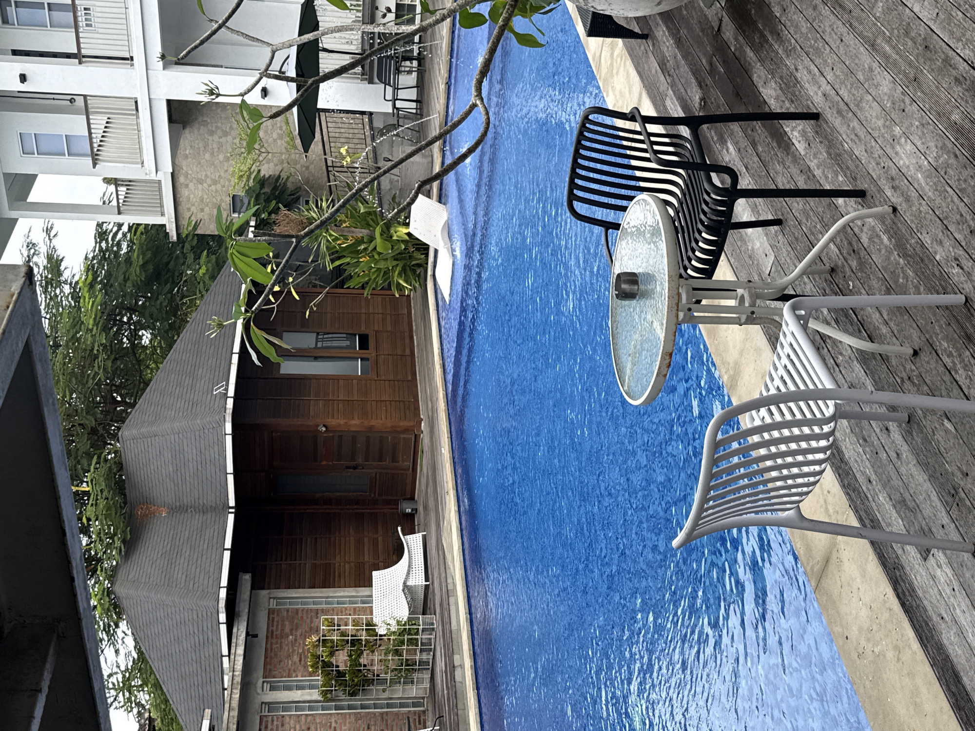 The image depicts a serene outdoor swimming pool area at a resort, likely located in Kuta Selatan, Indonesia, given the tropical flora and architecture. In the foreground, a bright blue swimming pool occupies the majority of the frame, with its surface rippled by gentle movement. To the left of the pool, a wooden deck runs alongside, furnished with two chairs and a small circular table. One chair is white and modern in design, while the other is black and more traditional. The table has a glass top and an ashtray. Beyond the pool, a white lounge chair is partially submerged in the water. In the background, a dark-tiled, pitched-roof building with wooden siding is visible, alongside a multi-story white building with balconies. Lush green trees and foliage surround the area, suggesting a warm, tropical climate. The lighting suggests it is daytime, possibly overcast. There are no people visible in the image.