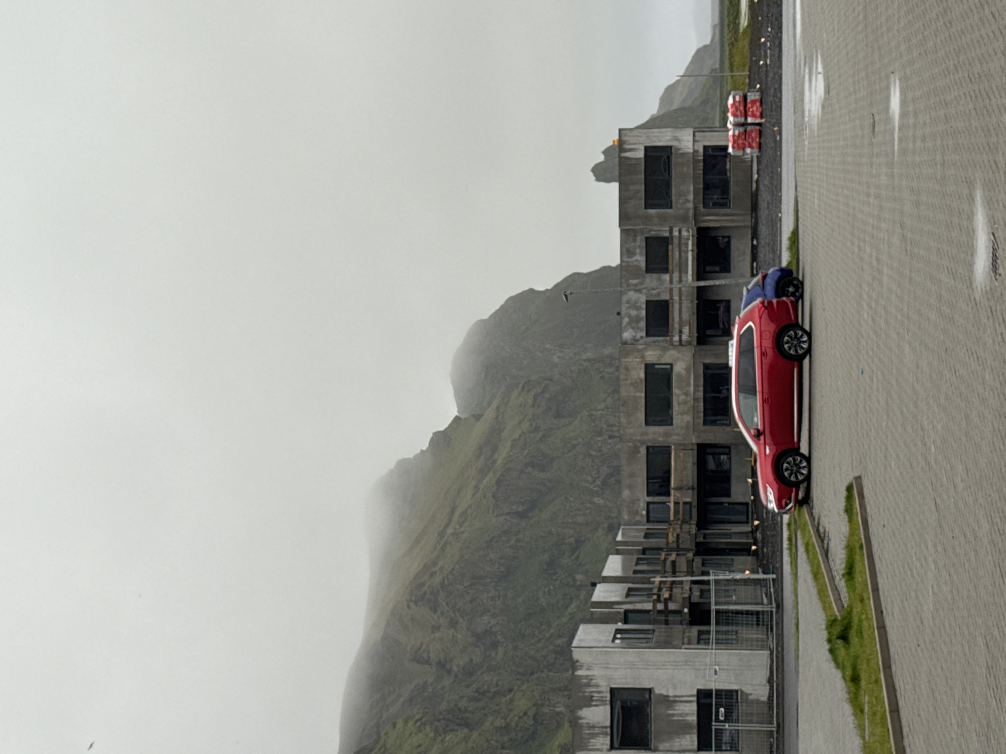 The image depicts a scene in Vik, Iceland, characterized by overcast weather and a mountainous backdrop partially obscured by fog. A modern, unfinished concrete building with multiple stories and rectangular window openings stands adjacent to a paved area where a red car is parked. Further along the pavement, stacks of red and white packaged goods are visible. The environment appears wet, with reflections suggesting recent rainfall on the pavement.