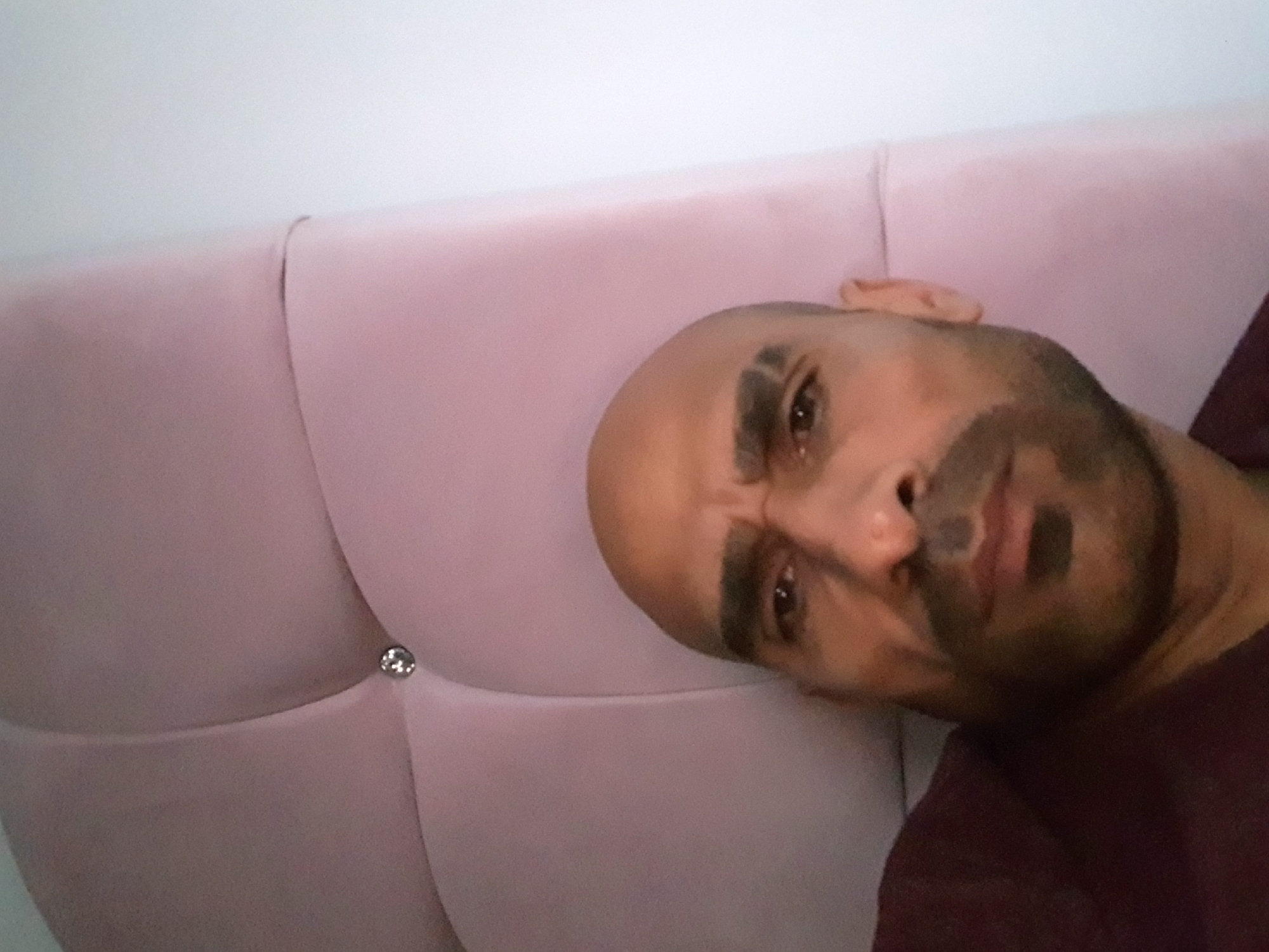 The image depicts a bald man with dark hair on his chin and a furrowed brow. He is lying on a pink, tufted headboard. A single button is visible on the headboard. The man is wearing a burgundy-colored shirt. The background consists of a plain white wall. The location is not discernible from the image.
