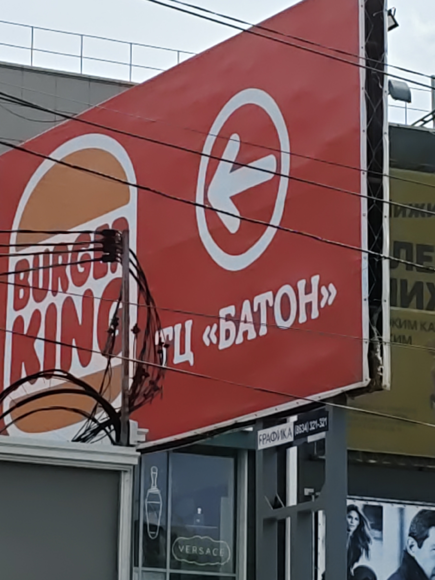 The image shows a close-up of a Burger King sign with the company logo and text in Cyrillic. The sign is red with white lettering and has an orange and white Burger King logo. The text on the sign appears to say "BURGER KING" in English and then something in Cyrillic, possibly "БАТОН" which translates to "baton." There are also power lines obscuring part of the sign. Below the Burger King sign, there is a smaller sign with Cyrillic text that reads "ГРАФИКА" and a phone number "(834) 321-321". To the right of this, a billboard with images of people is visible, and a sign that appears to say "VERSACE". The Burger King sign is on a building in what appears to be a commercial area. Based on the Cyrillic text and the Burger King branding, this image is likely from Novocherkassk, Russia.