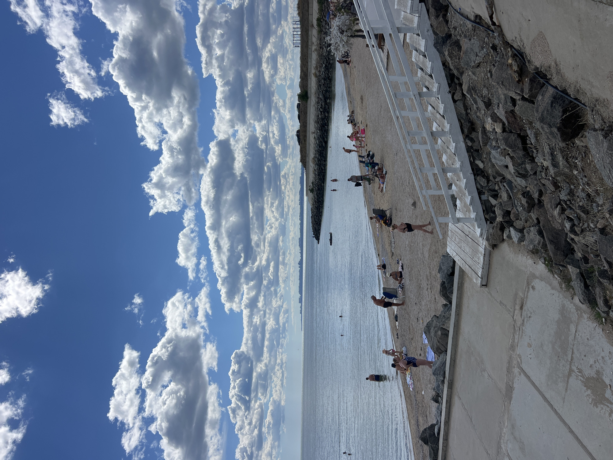 This is an eye-level, wide-angle photograph of a beach scene in Fontanka, Ukraine. The sky is bright blue with scattered white cumulus clouds. The sea is calm and stretches to the horizon where a distant city skyline is visible. The beach is composed of sand and pebbles, with a rocky embankment to the right of the frame.
Several people are on the beach, engaged in various activities. Some are in the water, either swimming or wading. Others are sunbathing on towels or chairs, or walking along the shore. There are also a few individuals standing and talking. A white wooden staircase leads down to the beach from the upper right of the frame.
The overall atmosphere is one of a pleasant summer day at the beach.