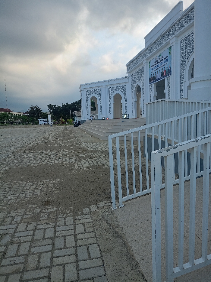 This image depicts the exterior of a large, modern white building, likely a mosque or community center, located in Meureudu, Indonesia, under an overcast sky.
The scene is dominated by the building, positioned to the right and center, featuring a multi-tiered entrance with numerous wide concrete steps leading to an elevated platform. The building's facade is white, adorned with intricate dark grey decorative patterns, notably around a series of large arched openings. A prominent green and white banner, displaying text in an Indonesian language and a group photograph, is affixed to the building's upper facade, suggesting an event. Near the base of the steps on the elevated platform, several illuminated stage lights or similar equipment are visible.
In the immediate foreground, a white metal railing curves into the right side of the frame, leading to a concrete ramp or pathway. The ground in front of the building is a paved area constructed from rectangular grey interlocking bricks, interspersed with patches of light brown dirt. To the left of the building, a few motorcycles or scooters are parked on this paved surface.
Further into the background on the left, an area of green foliage and trees is visible, with distant low-rise buildings featuring red roofs. A tall, slender communication or transmission tower extends vertically against the cloudy sky on the far left horizon. The sky itself is heavily overcast with dense grey cloud cover. No individuals are clearly discernible within the frame.