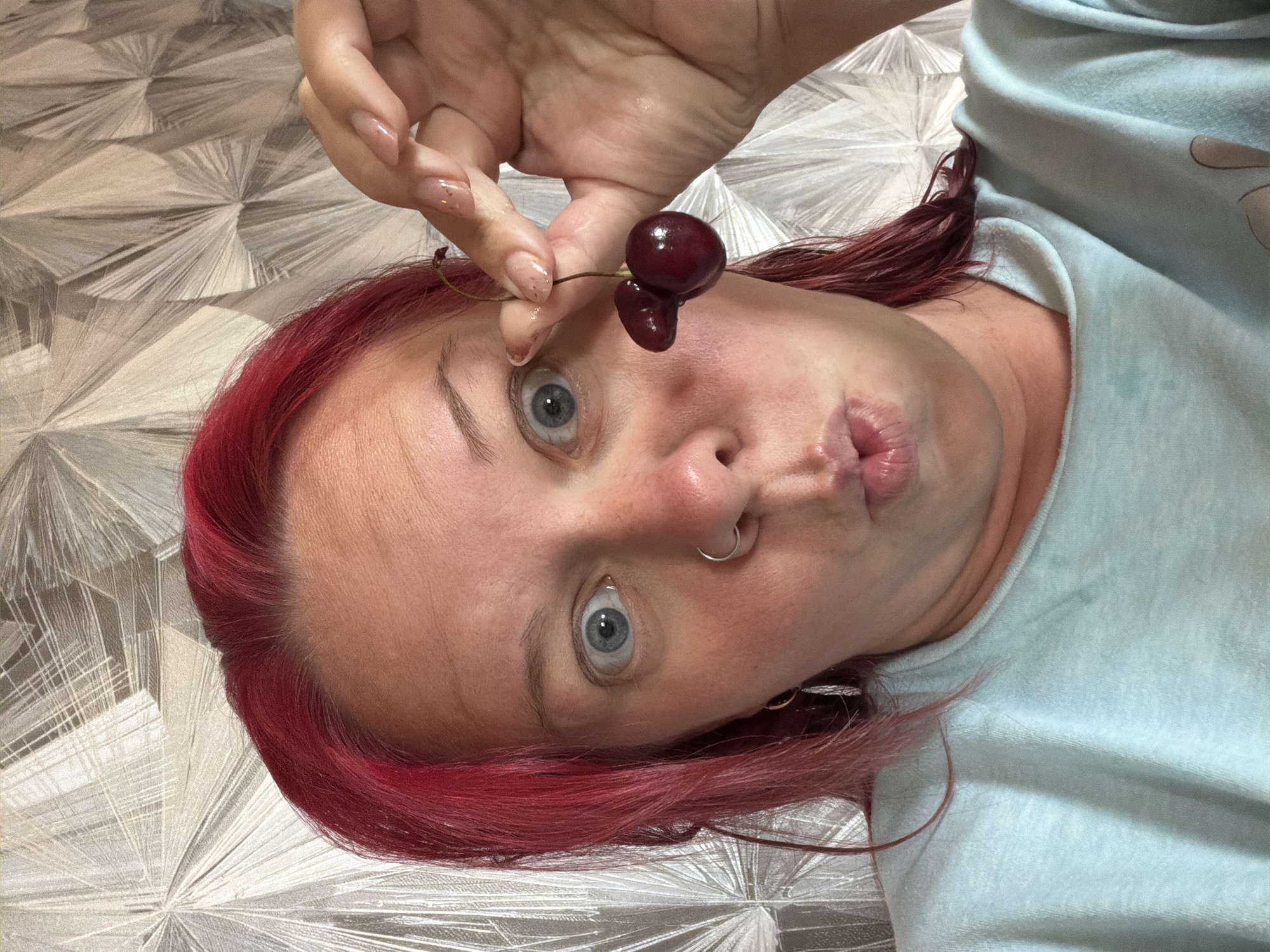 A close-up, low-angle selfie shows a person with bright pink hair, blue eyes, and a septum piercing. They are wearing a light blue sweatshirt and making a "pucker" face while holding up two cherries to the camera. The background is a grey and white patterned wallpaper. The individual's skin appears flushed. The scene does not provide any information about the location in Chaikovsky, Russia.