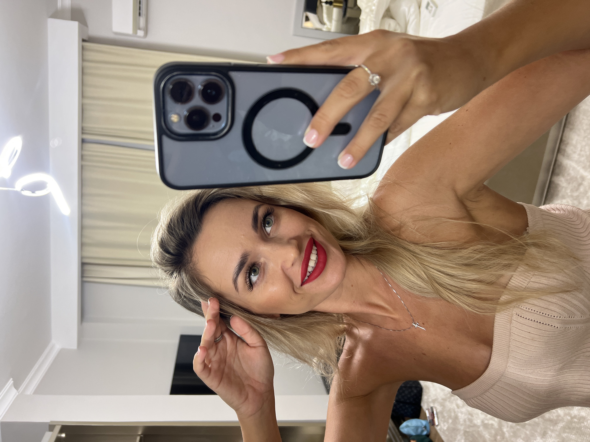 A blonde woman with green eyes and red lipstick is taking a selfie in a bedroom. She is wearing a light-colored, strapless top and a silver cross necklace. Her right hand is touching her hair, and her left hand is holding a smartphone, showing a ring on her left ring finger. The room appears to be well-lit with overhead lighting. In the background, there are curtains, a closet, and a bed. The image does not contain any specific visual cues to indicate the location is Sharm El Sheikh, Egypt.