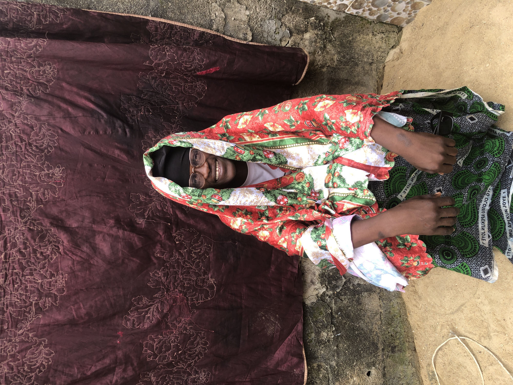 A person wearing glasses and a headscarf with a Christmas-themed pattern is kneeling in front of a dark red fabric with floral embroidery. The person is smiling and looking at the camera. The background consists of a textured concrete wall and a sandy ground. A white rope is visible on the ground to the left. The scene appears to be outdoors in Ado, Nigeria.