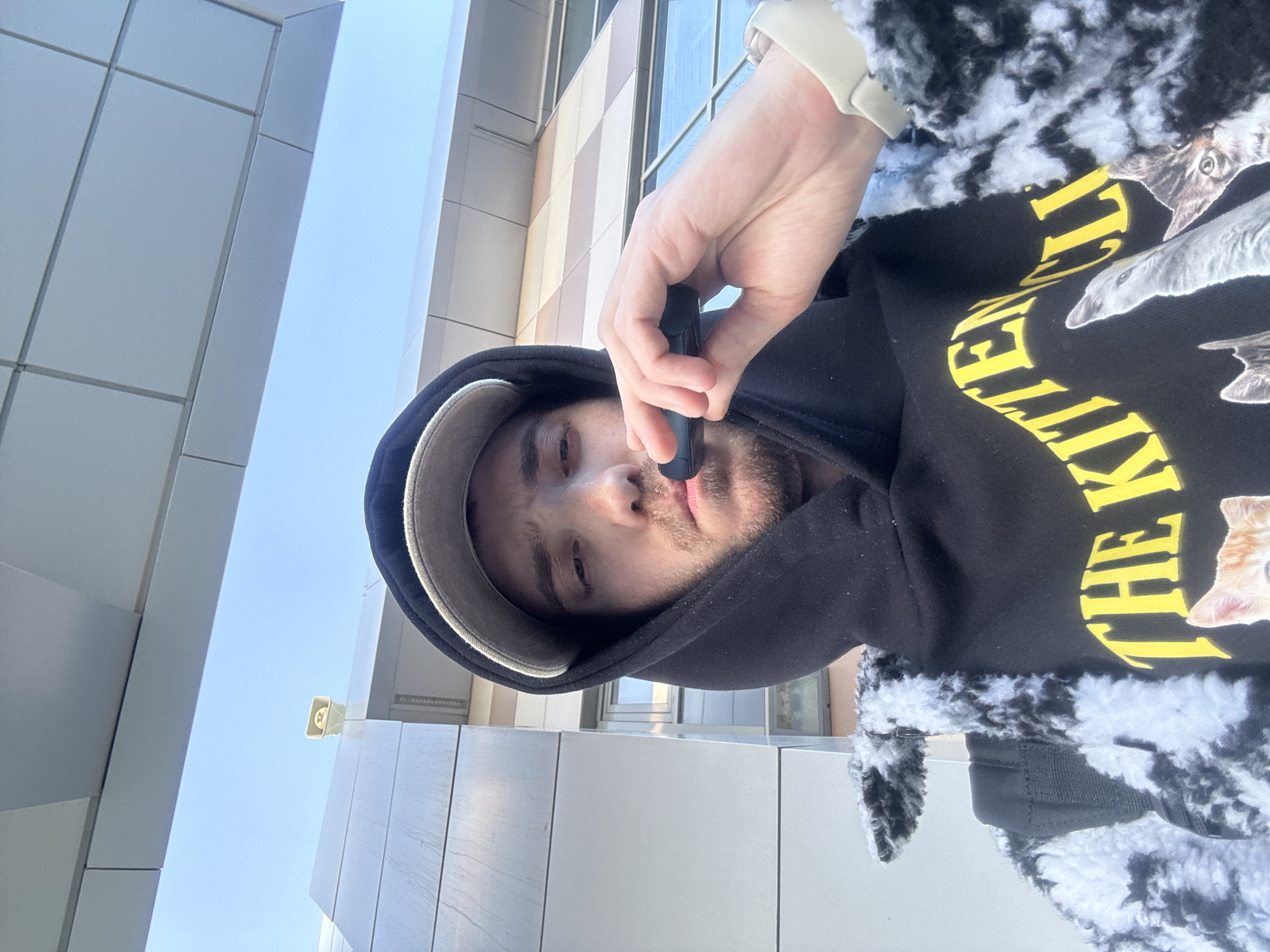 A low-angle, outdoor photograph depicts a person holding a vaping device to their lips. The individual is wearing a black hoodie with a cat print and yellow lettering that reads "The Kitten." The hoodie is partially covered by a black and white fuzzy jacket. The person also wears a baseball cap. The background features a modern building facade with horizontal panels and a clear blue sky. Based on visual references, the location in the city of Bakchasarai, Russia, cannot be confirmed.