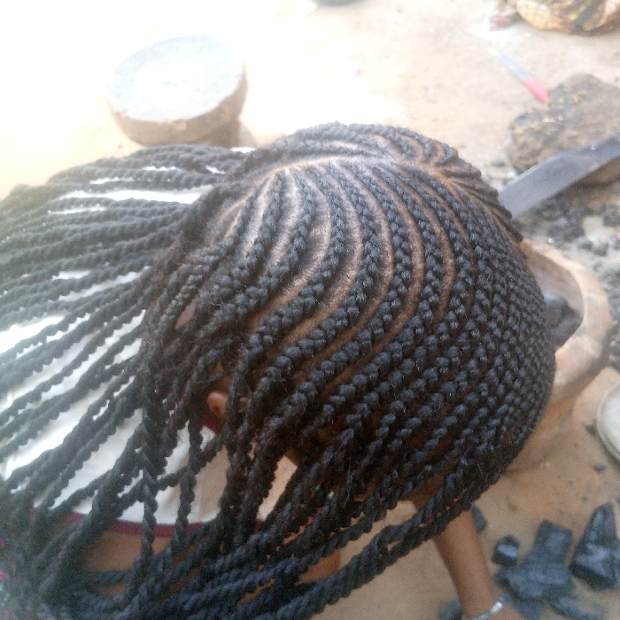 An overhead view captures the head of a person in Ibreda, Nigeria, featuring an elaborate hairstyle composed of intricate black cornrow braids that curve across the scalp, and numerous long, twisted hair extensions that hang downwards over the left shoulder. The person's left shoulder is covered by a white garment with red trim. A silver-colored bracelet is visible on their right wrist. The outdoor setting includes a light-colored ground surface, appearing to be dirt or concrete. In the background, objects on the ground include a circular flat stone, a dark wooden mortar or similar large object, several dark, irregularly shaped lumps (consistent with charcoal), a red-capped pen, and a dark elongated object.