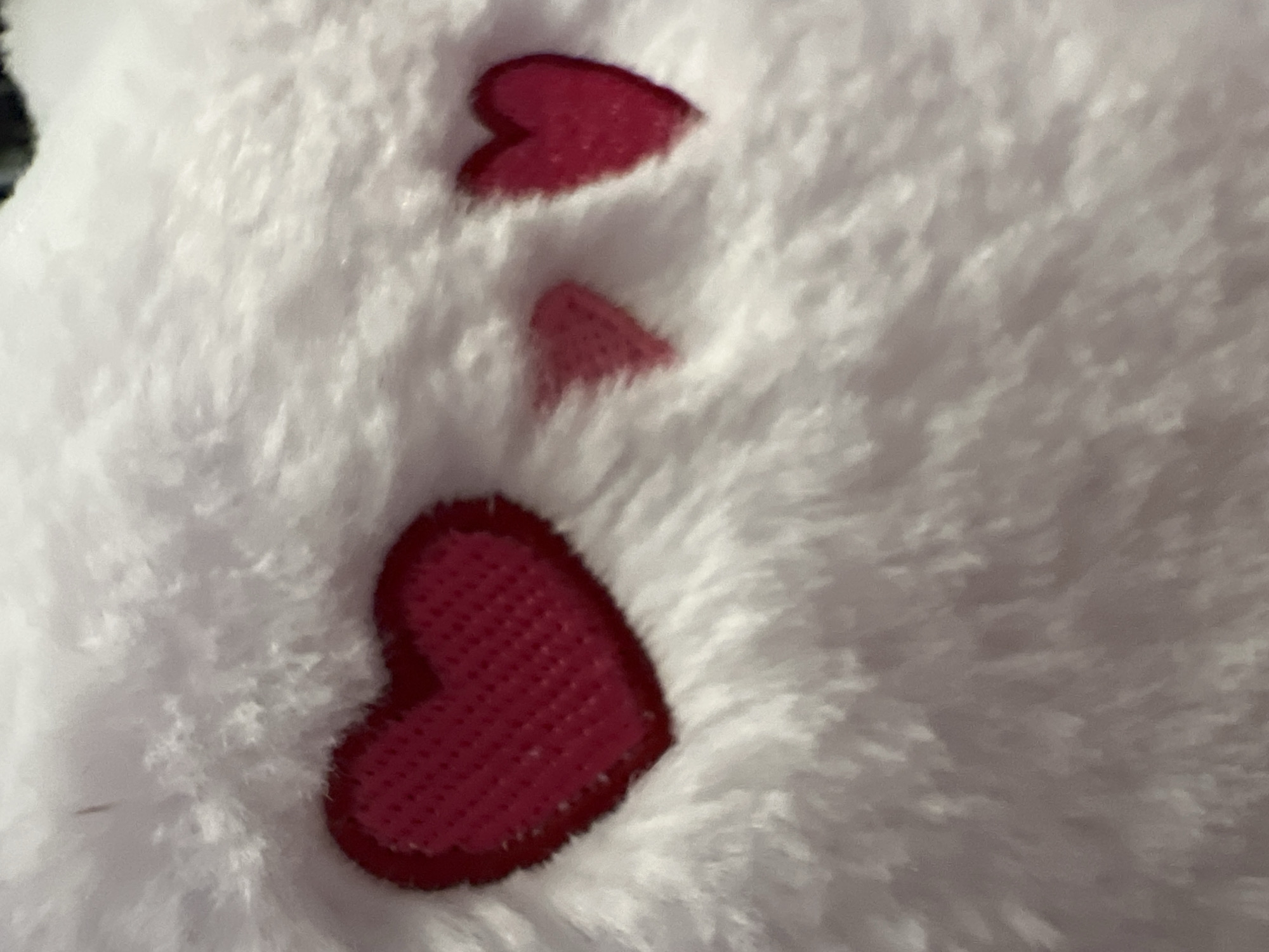 A close-up shot shows a white, fluffy material with three red heart-shaped patches of varying sizes sewn onto it. The largest heart is at the bottom, with a smaller heart above it and an even smaller piece that could be part of a heart above that.
The image does not appear to be taken in Moscow, Russia and the scene shows no people, subjects, objects, actions, interactions, or the environment.