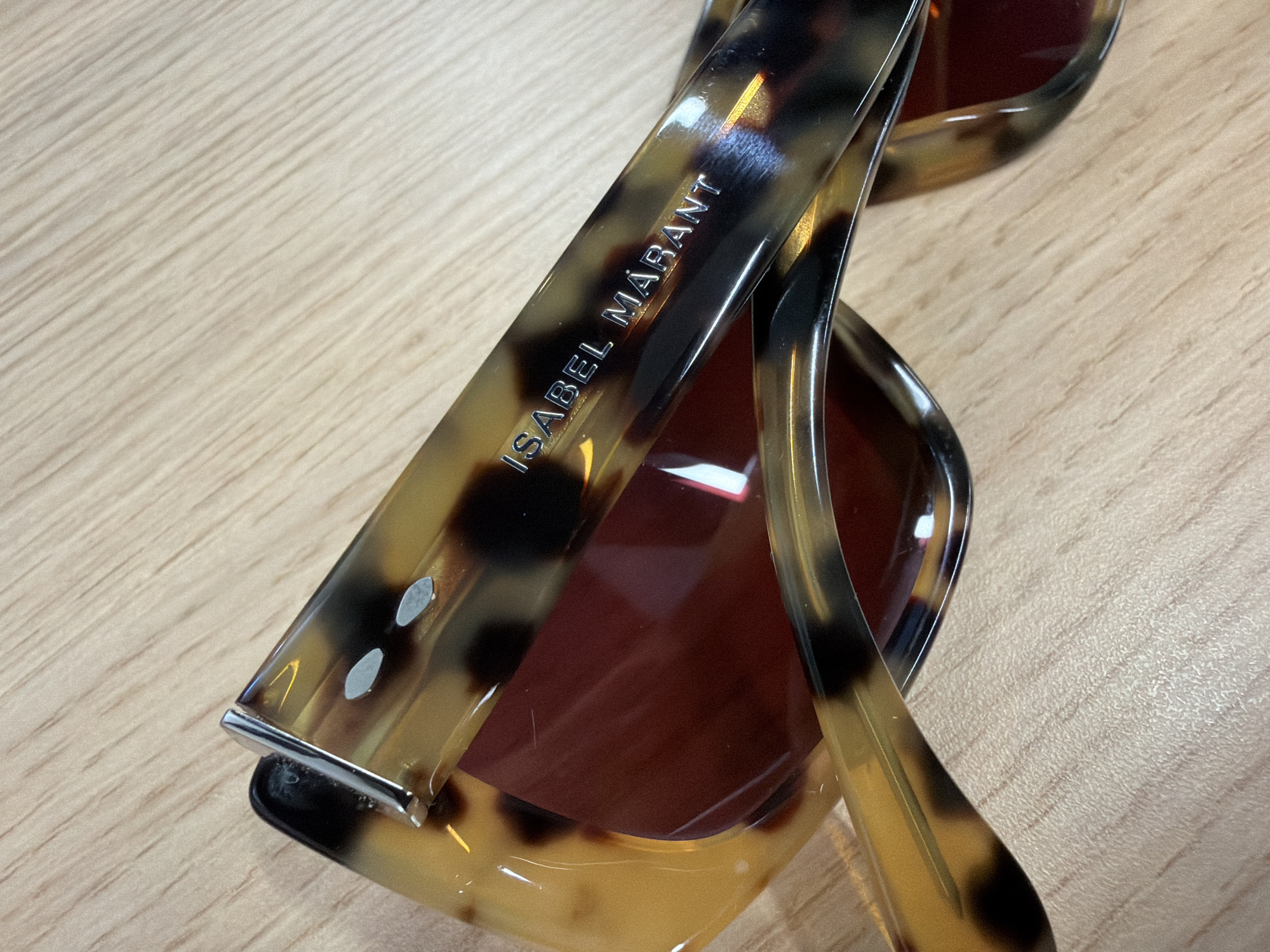 The image shows a pair of Isabel Marant sunglasses with a tortoise shell pattern. The sunglasses are resting on a light-colored wooden surface. The focus is on the left arm of the glasses, where the brand name "ISABEL MARANT" is visible. The location is not discernible from the image.