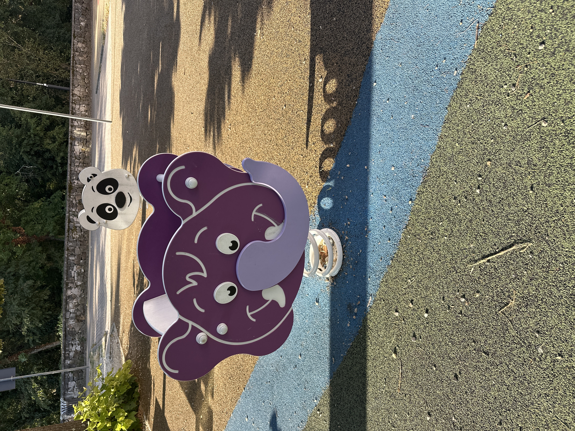 In a park in Trieste, Italy, a brightly colored children's playground is illuminated by the sun. In the foreground, a purple elephant-shaped spring rider is centrally positioned. Its large, friendly face, complete with wide eyes and a smiling trunk, is rendered in a cartoonish style. The elephant is mounted on a white spring, which is secured to the ground. Behind the elephant, slightly out of focus, is a white panda-shaped spring rider, its black and white markings distinct against the sunny background. The playground surface is a mosaic of textures and colors, with sections of tan, blue, and green rubberized material. Shadows of trees and playground equipment are cast across the surface, indicating a sunny day. In the background, a stone wall and lush green trees suggest a natural, park-like setting. A few small pebbles and pieces of debris are scattered across the playground surface.