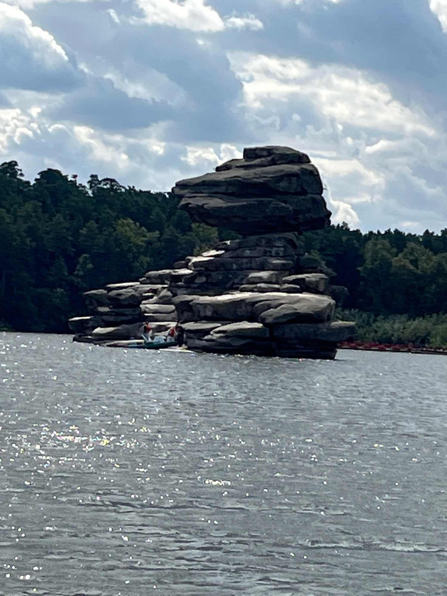 A wide shot captures a large rock formation jutting out of a body of water. The formation consists of several stacked, flat rocks, with the topmost rock being significantly larger and overhanging the lower layers. A small, green boat is visible near the base of the rock formation, with two individuals in red shirts and one person in a blue shirt onboard. The water is choppy and reflects sunlight, creating a sparkling effect. In the background, a dense forest of dark green trees lines the horizon, backed by a cloudy sky with patches of blue. The scene appears to be a natural landscape.