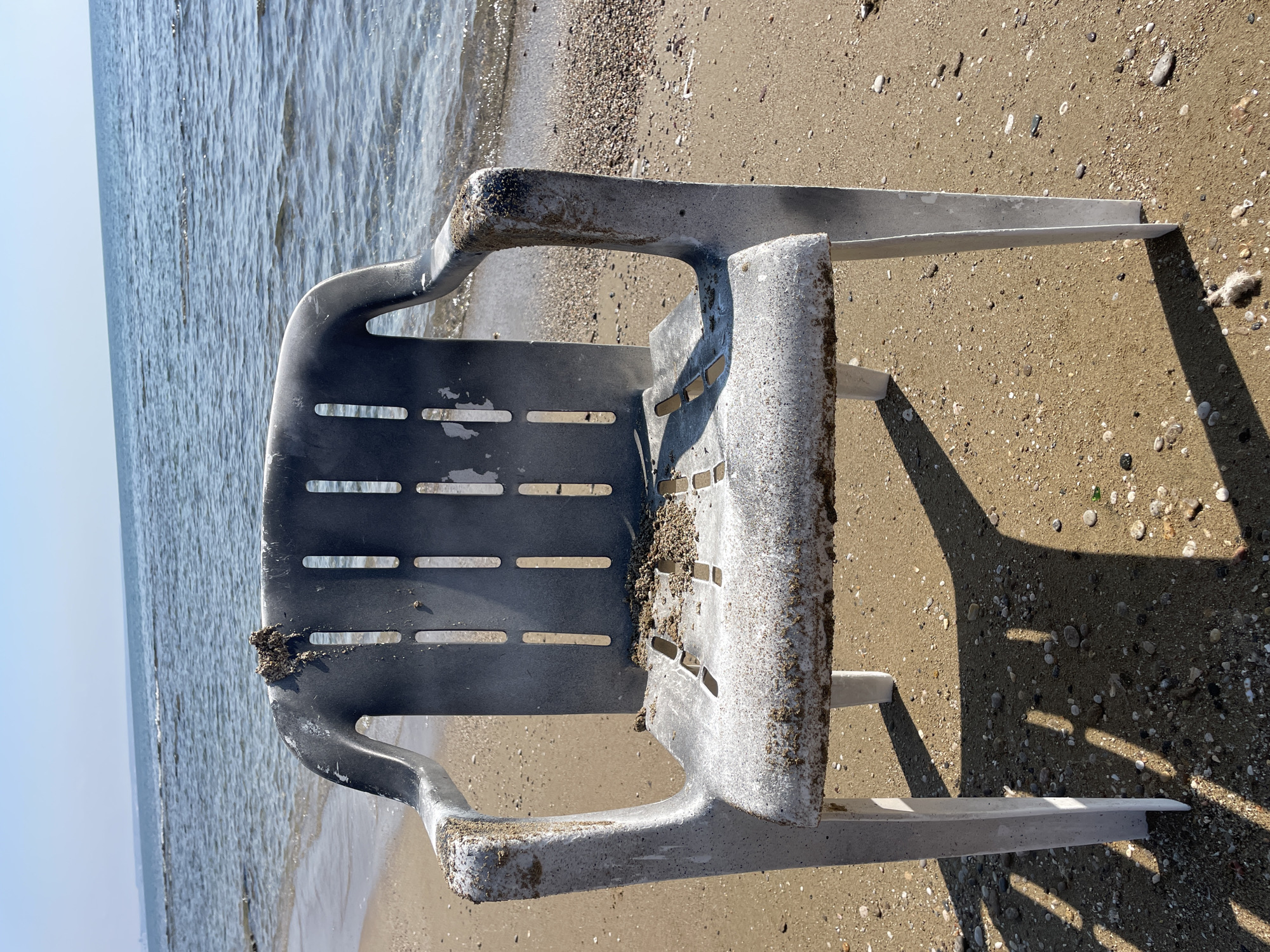 An abandoned plastic chair sits on a sandy beach, facing the sea. The chair is weathered and covered in sand and what appears to be dried seaweed. The ocean is calm with gentle waves lapping the shore. The sky is clear and blue. The location is the beach in Agios Sergios, Northern Cyprus.