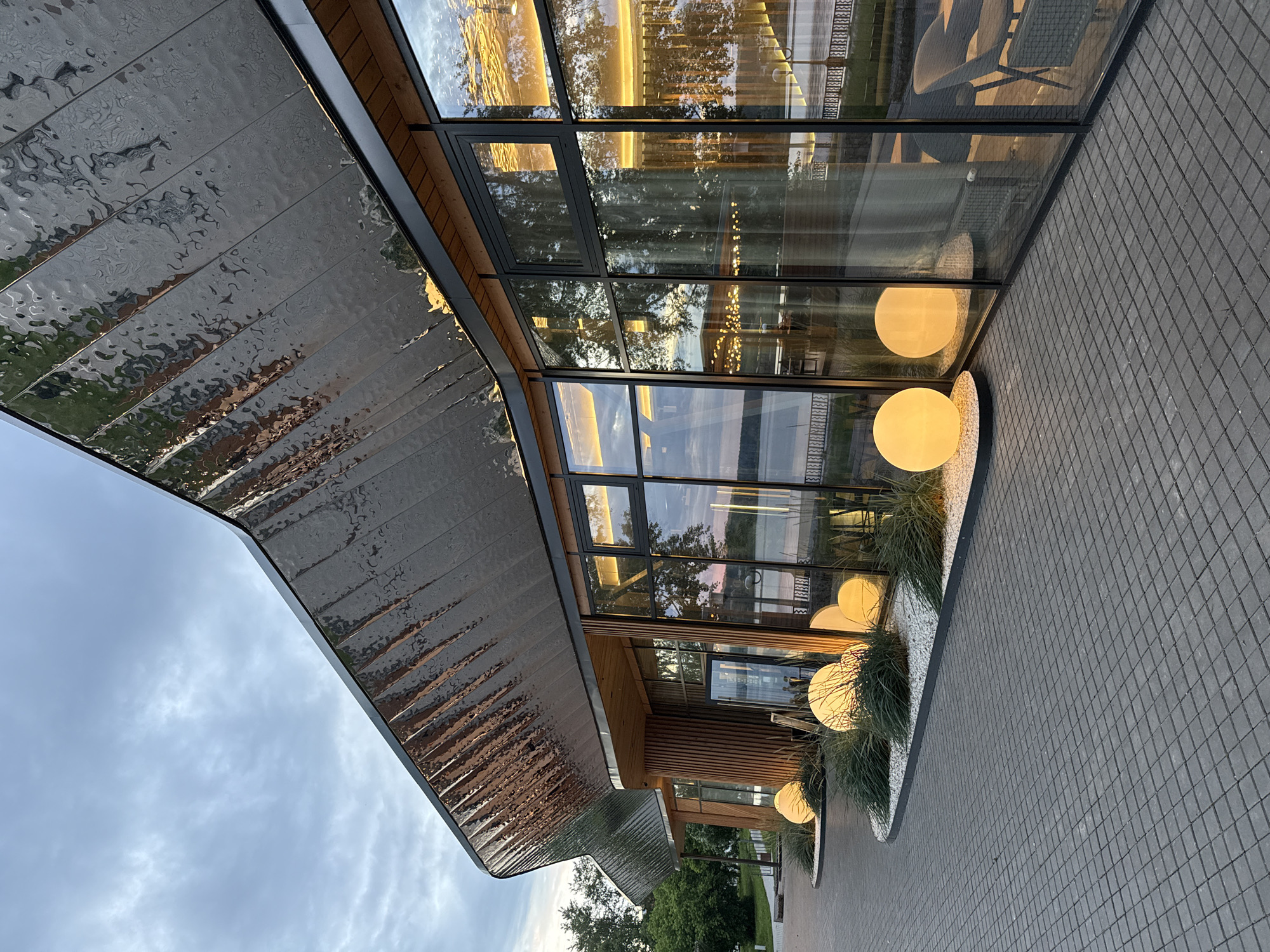 The image shows the exterior of a modern building with a distinctive, textured metallic roof. Large glass windows reflect the surrounding environment, including trees and the sky. Inside the building, warm lighting is visible, with some string lights and larger spherical lamps illuminating the interior.
Outside the building, a paved walkway made of dark gray square tiles runs alongside a landscaped area. This area features lush green grass and several large, spherical outdoor lamps emitting a soft yellow glow. These lamps are interspersed with white gravel and plants, creating an inviting ambiance.
The scene appears to be set during the late afternoon or early evening, as indicated by the warm, golden light reflecting in the windows and the soft glow of the lamps. The sky is partly cloudy, suggesting mild weather. The architectural style of the building, with its blend of natural wood elements and sleek glass and metal, points to a contemporary design. The location context indicates this is Votkinsk, Russia. There is no visible text in the image. The overall mood conveyed is one of modern elegance and tranquil evening illumination.