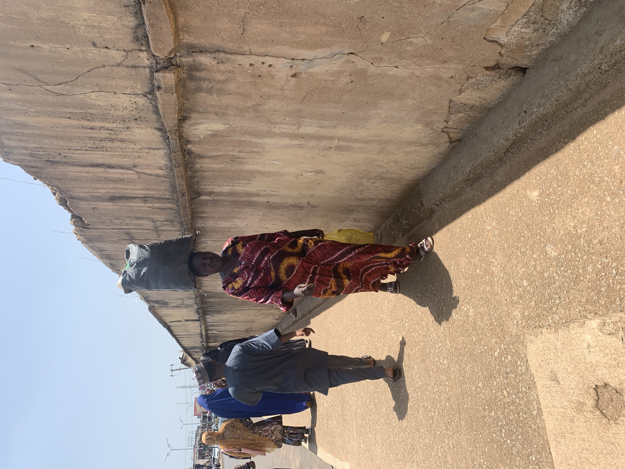 In Jos, Nigeria, a woman walks down a sidewalk carrying a large black trash bag on her head. She is wearing a colorful, patterned dress and sandals. To her left, a man in a gray outfit and a patterned hat walks away from the camera. Behind him, other people in traditional clothing are visible, including a woman in a blue hijab and another in a gold and brown outfit. The sidewalk is paved and a weathered concrete wall runs along the right side of the image. The environment appears to be an urban street scene with a bright, clear sky.
