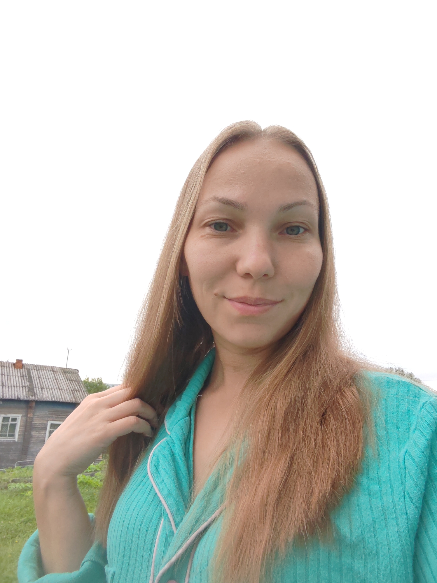 A woman with long blonde hair is taking a selfie outdoors. She is wearing a turquoise-colored pajama top with white trim. Her left hand is touching her hair. In the background, there is a wooden house with a gray roof and a window. The area around the house appears to be a yard with green grass and some foliage. The sky is overcast. This scene is located in the city of Chuchlem, Russia.