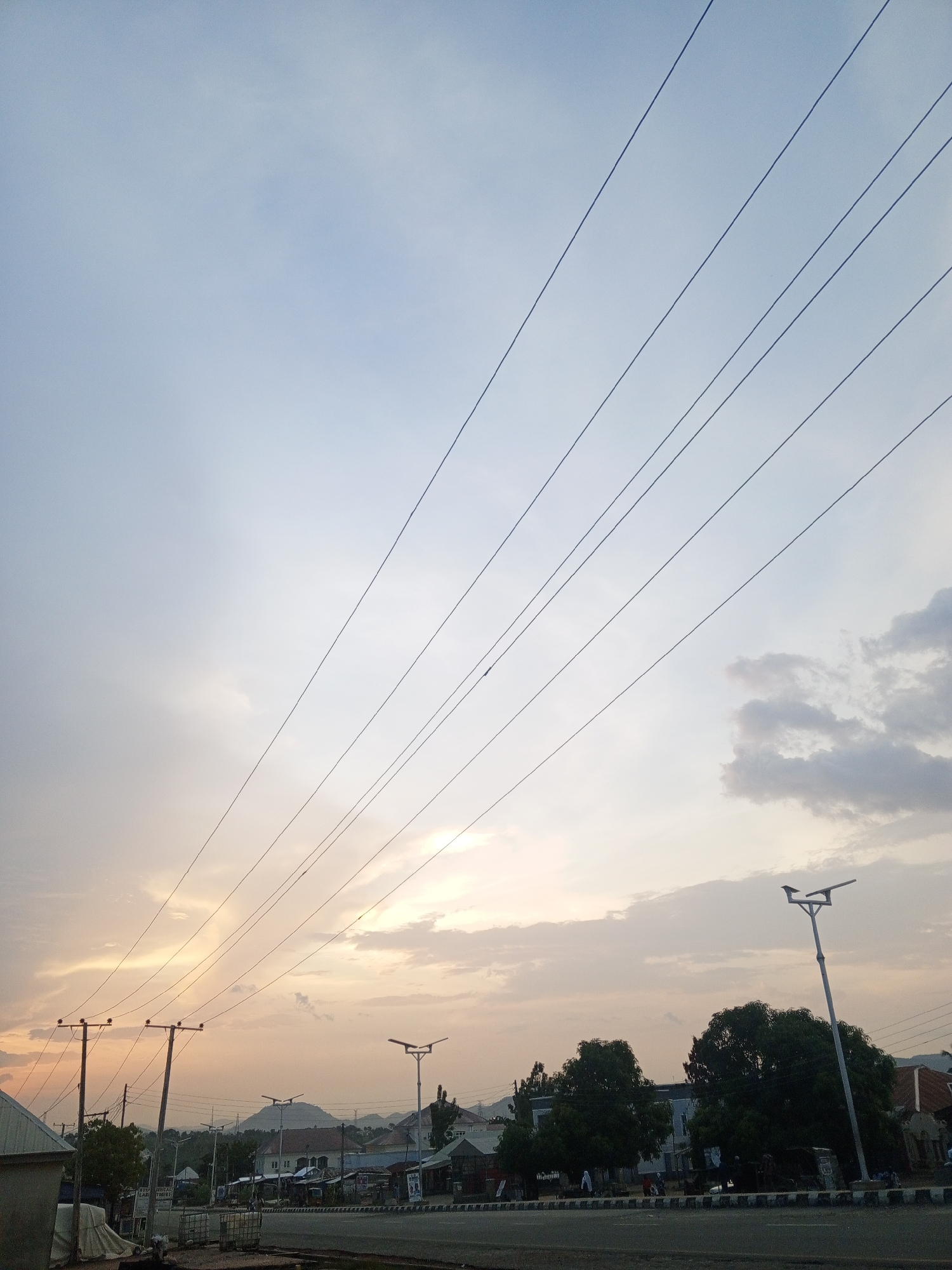 The image shows a street scene in an urban area, with an expanse of sky above. Overhead power lines stretch diagonally across the frame. The sky is a mix of light blue and orange hues, suggesting either sunrise or sunset. Below the power lines, there are buildings, trees, and street lights lining the road. A road with a black and white curb runs along the bottom of the image. The location is Unknown, Unknown. There are no people clearly visible in the image.