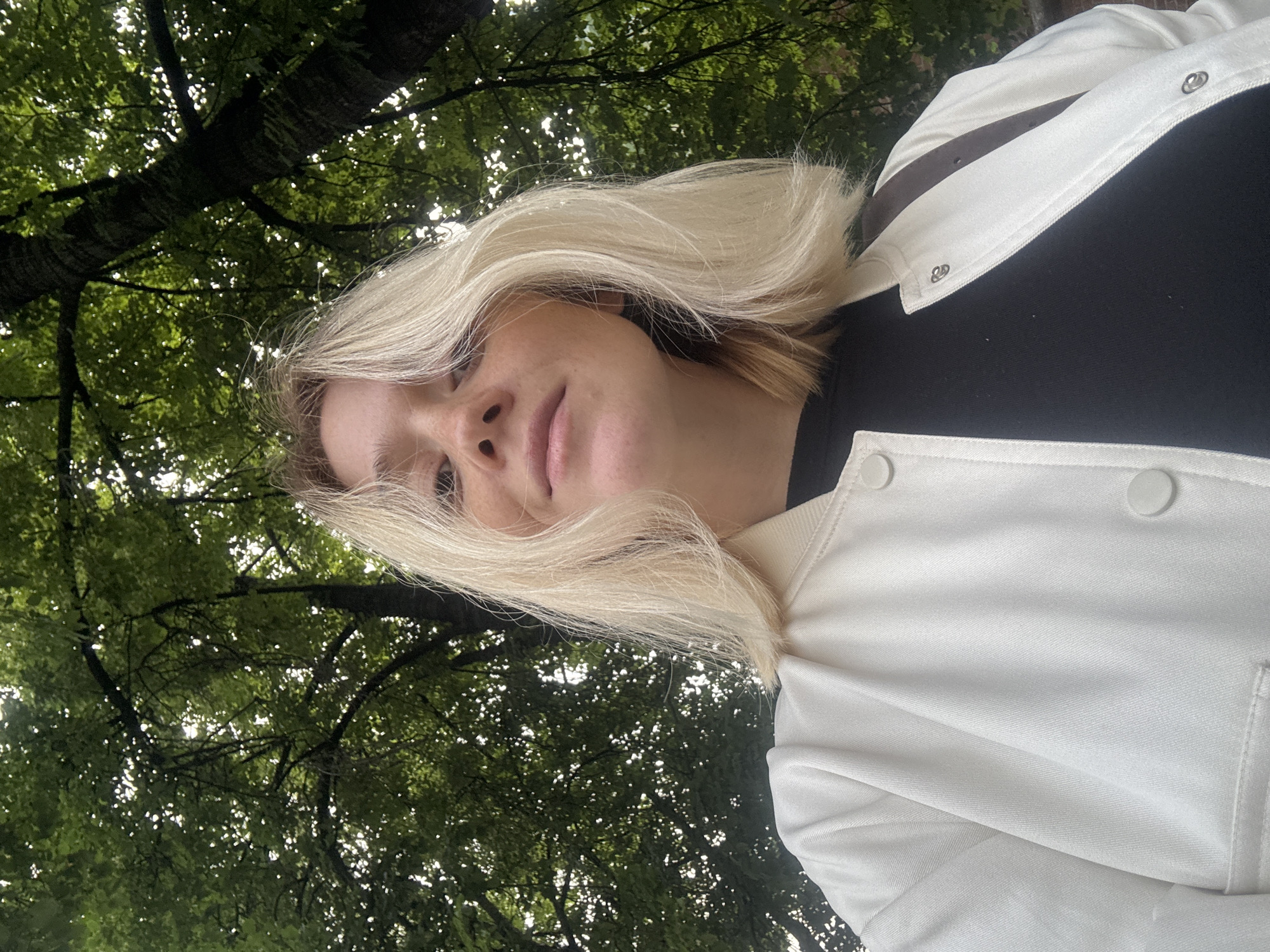 In an outdoor, low-angle shot, a young woman with blonde hair and freckles looks up at the camera. She is wearing a white bomber jacket over a black t-shirt. The background is dominated by the dense, green foliage of trees, suggesting a park or a wooded area within the city. The lighting is natural, with dappled sunlight filtering through the leaves. No specific landmarks of Nizhny Novgorod are visible.