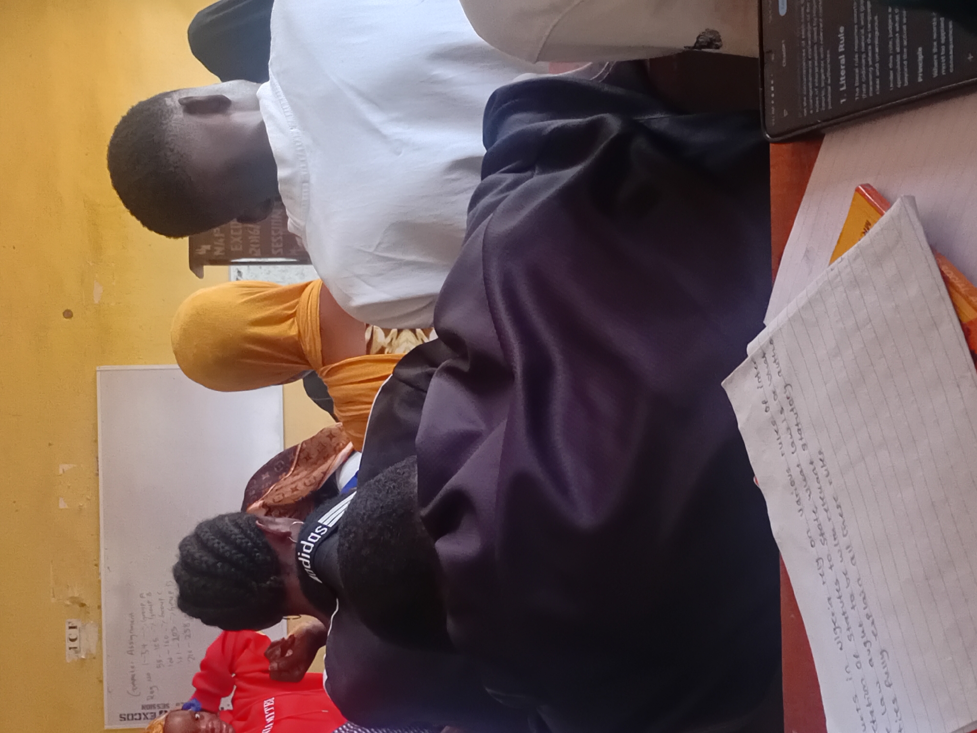 The image depicts an indoor scene, likely a classroom or lecture hall, with several individuals present. The setting appears to be in Ogbogbo, Nigeria, as indicated by the whiteboard text and a handwritten note, though this cannot be definitively confirmed solely from the visual information.
In the foreground, a person wearing a black hoodie with "adidas" branding on the collar is partially visible, facing away from the camera. To their right, a person in a white shirt is also seen from behind. In the middle ground, a person wearing a yellow hijab is seated. To the left, a person in a red hoodie is facing the camera, touching their hair.
A whiteboard is visible in the background, with handwritten text in English, including what appears to be an assignment list with registration numbers and group assignments. Another section of text on the whiteboard seems to be related to "NMAP EXEC 2016 SESSION".
A handwritten note on lined paper, also in English, is placed on a red surface in the lower part of the frame. The text on this note discusses rules of interpretation of statutes in Nigeria.
A smartphone is visible on the right side of the frame, displaying text that appears to be from a digital document, possibly related to legal principles. The environment is characterized by yellow walls and artificial lighting. The overall atmosphere suggests an academic or educational setting.