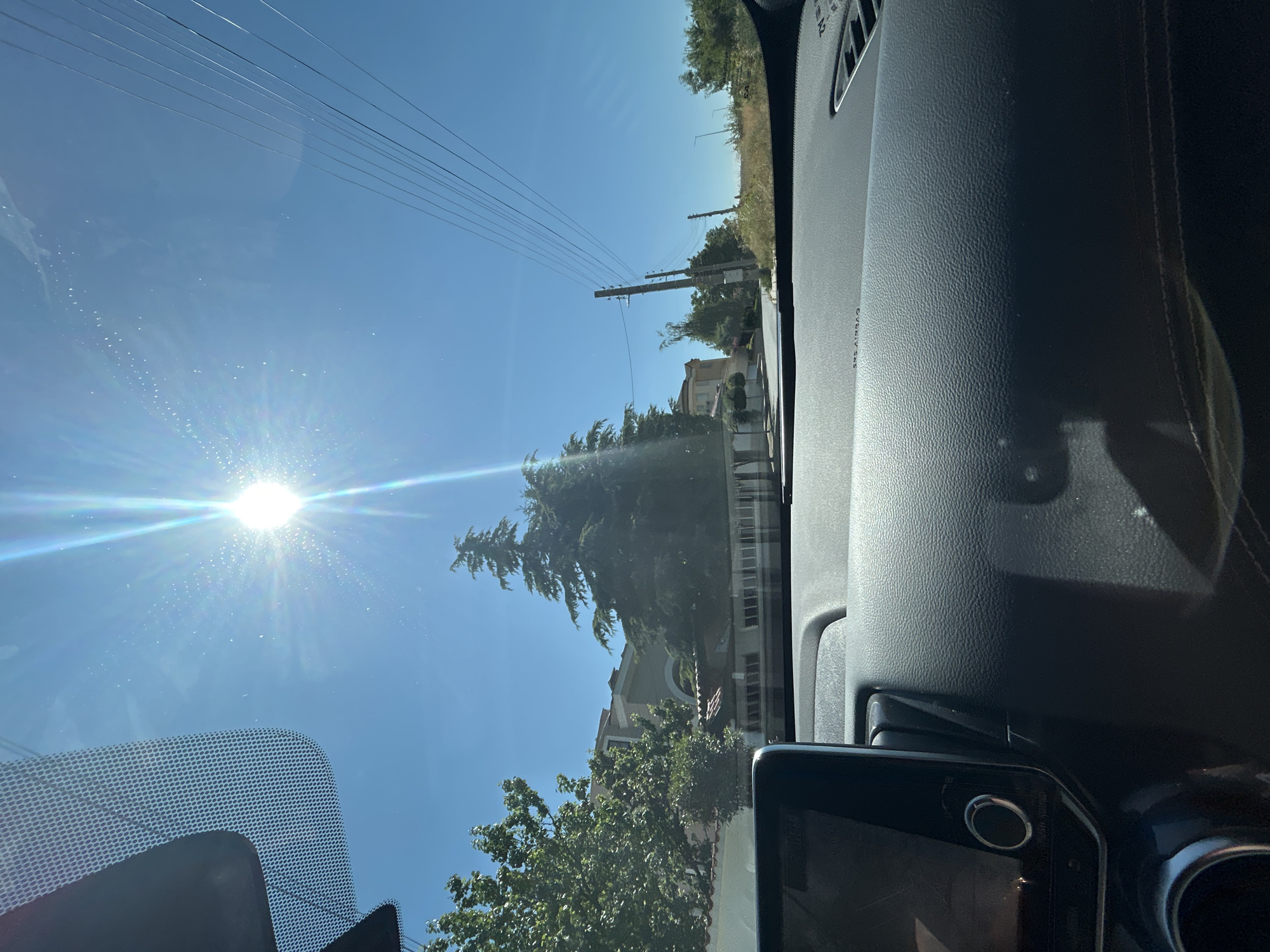 A sunny daytime view from inside a car in Markovo, Bulgaria, shows a clear blue sky with the sun prominently shining, creating lens flares. The foreground features the car's dashboard, including a touchscreen display on the left and textured plastic surfaces. In the midground, a street lined with trees and houses is visible. Two tall coniferous trees stand out, with a light-colored building behind them. Utility poles and power lines run along the right side of the road. The environment appears to be a residential area with well-maintained vegetation.