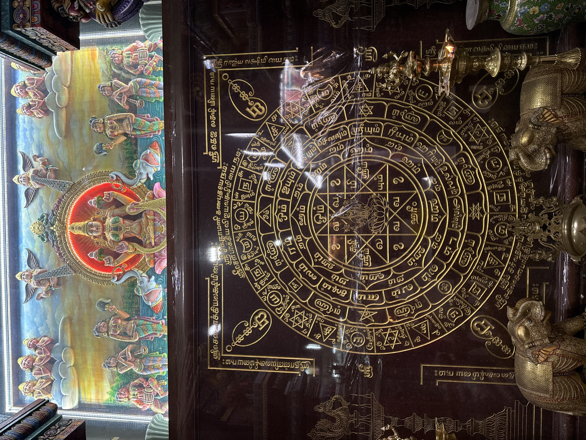 The image shows a display within a Buddhist temple in Bangkok, Thailand. On the left, there is a three-dimensional artwork depicting Hindu deities and celestial figures with a central female deity surrounded by an illuminated halo. To the right is a dark wooden panel with intricate gold inscriptions and symbolic designs. A complex mandala is etched on the wood, surrounded by Tamil text and various Hindu symbols. The panel is also partially obscured by a protective plastic covering. Gilded ornamental objects, including a lampstand and possibly figures of elephants, are positioned near the panel. The general environment suggests a sacred and ornate space within a religious building.