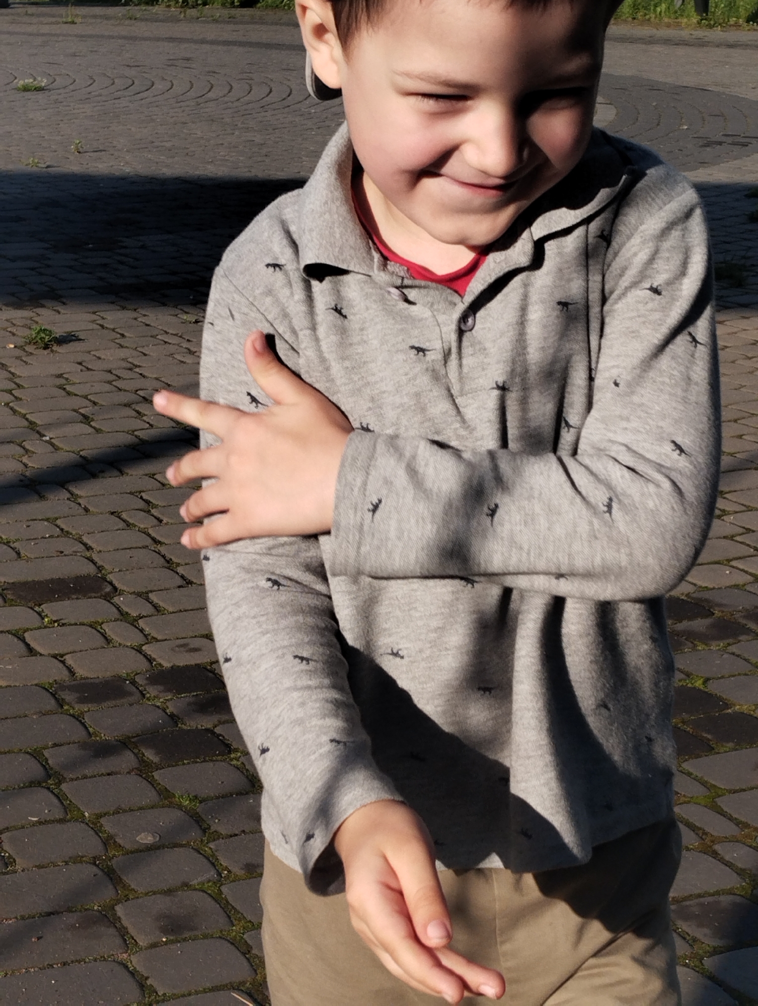 In an outdoor, paved area, a young boy with dark hair and a light complexion is captured from the chest up. He wears a light gray long-sleeved shirt with a pattern of dark silhouettes, possibly animals, and a red undershirt visible at the collar. His right arm is bent and rests across his chest, while his left hand is raised with fingers extended. The boy is looking down and to his left with a slight smile. The background consists of dark gray cobblestones with some patches of green vegetation. Shadows are cast across the scene, indicating a sunny day. This setting is consistent with an urban environment, possibly a public square or walkway.