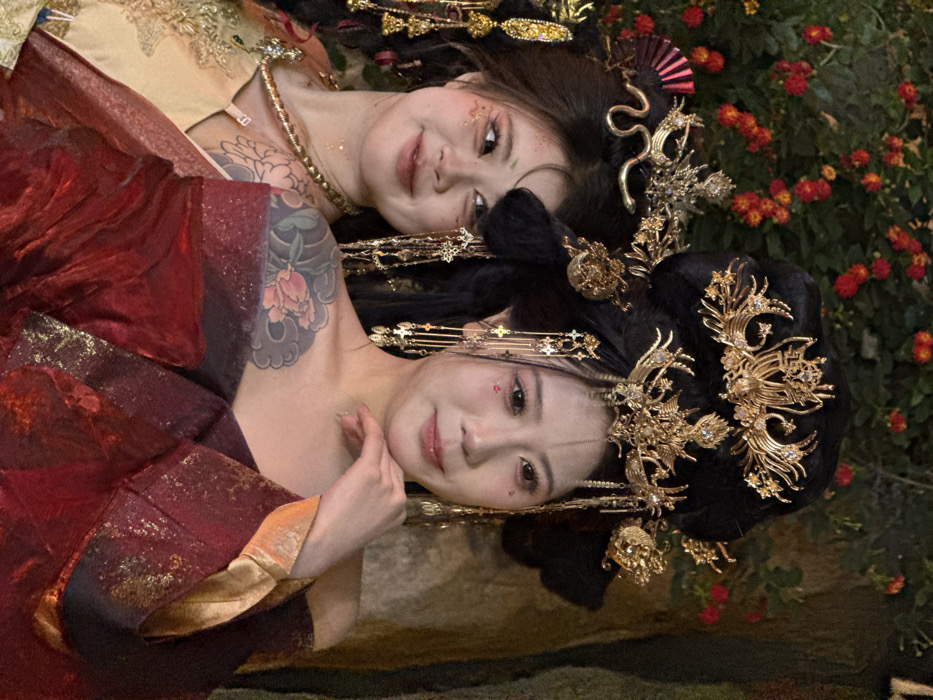 This image features two East Asian females, appearing to be in their 20s or 30s, posed in elaborate traditional Chinese-style attire. The scene is set against a dark, blurred background, consistent with an outdoor or themed indoor location, possibly in Yuzhong District, China. The background reveals green foliage with small red-orange flowers on the left and a darker, textured surface resembling rock or tree bark on the right.
The woman in the foreground, positioned prominently on the right, wears a deep red garment with visible gold trim. Her black hair is styled in an ornate updo adorned with multiple gold headpieces, including a central piece resembling a phoenix or mythical bird and numerous dangling gold chains with small cross-like charms. She has visible makeup with red accents below her eyes and a subtle vertical line painted on her nose bridge. A large, colorful floral tattoo is visible on her left shoulder and upper arm. She looks towards the viewer with a slight, contemplative smile, her right hand gently touching her chin.
Partially visible behind and to the left is the second woman, who wears a gold-colored traditional garment. Her dark hair is also elaborately styled with a gold headpiece featuring a fan-like element and a serpentine design, complemented by additional gold dangling ornaments. She displays makeup with iridescent glitter around her eyes and small green accents. A portion of a floral tattoo is visible on her upper right arm. She smiles softly while looking towards the viewer.
