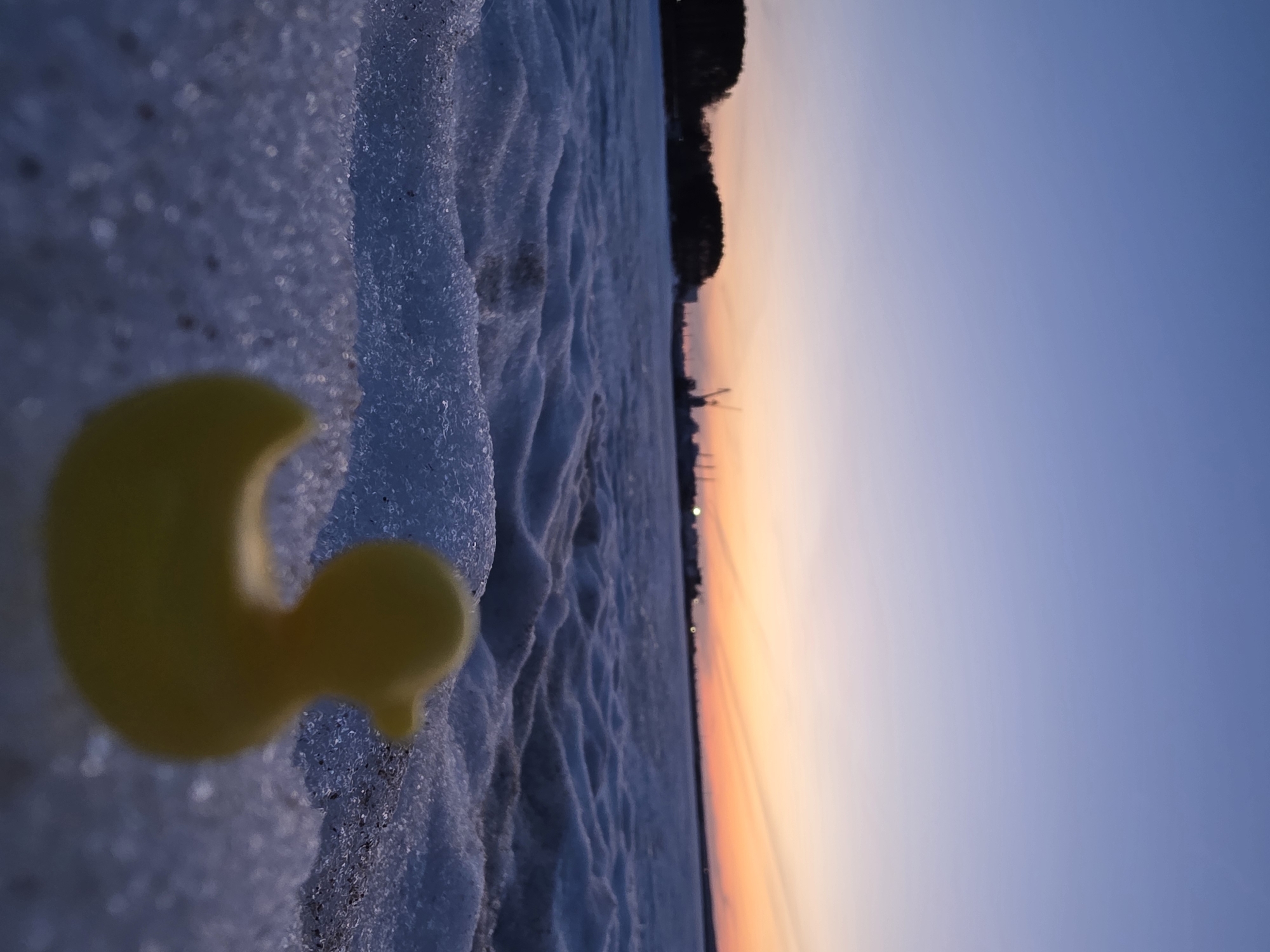 A close-up, ground-level, tilted shot depicts a yellow rubber duck partially visible in the left foreground, nestled among icy or snow-covered ground. The background reveals a horizon with a gradient of colors transitioning from orange to blue, suggestive of either sunrise or sunset. Further off in the background, faint silhouettes of what appear to be trees and possibly industrial structures can be discerned on the horizon, potentially indicating the city of Chaikovsky, Russia, where the image was reportedly taken. The ice or snow exhibits a textured surface, with varying depths and formations.