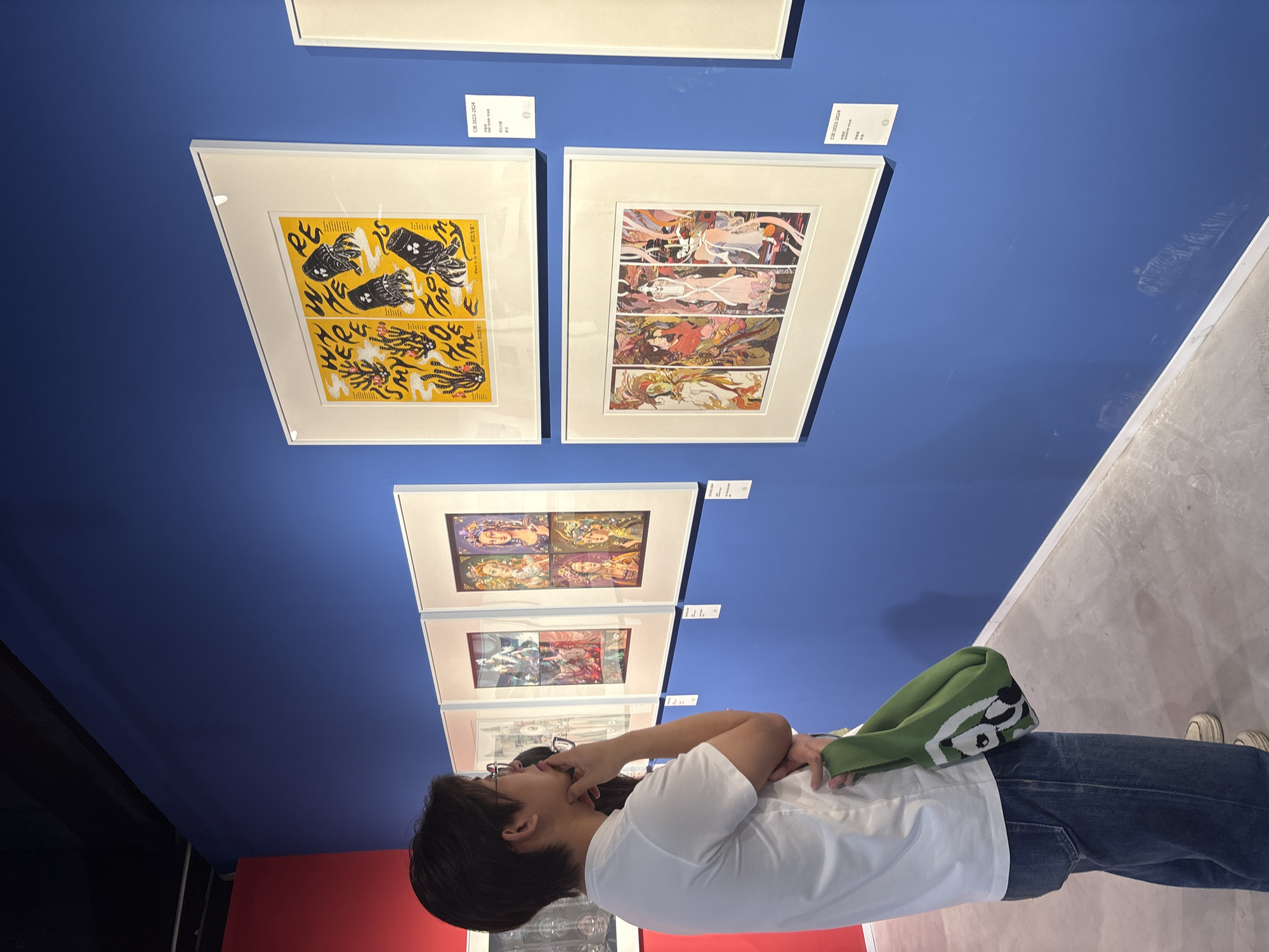 The image shows a person in an art gallery in Nan'an District, China. The person is wearing glasses, a white t-shirt, jeans, and white sneakers. They have a green bag over their shoulder and are looking at a series of framed artwork on the wall, each consisting of a four-panel comic strip with different art styles. The art gallery has a blue wall, a red wall in the corner, and a concrete floor.
