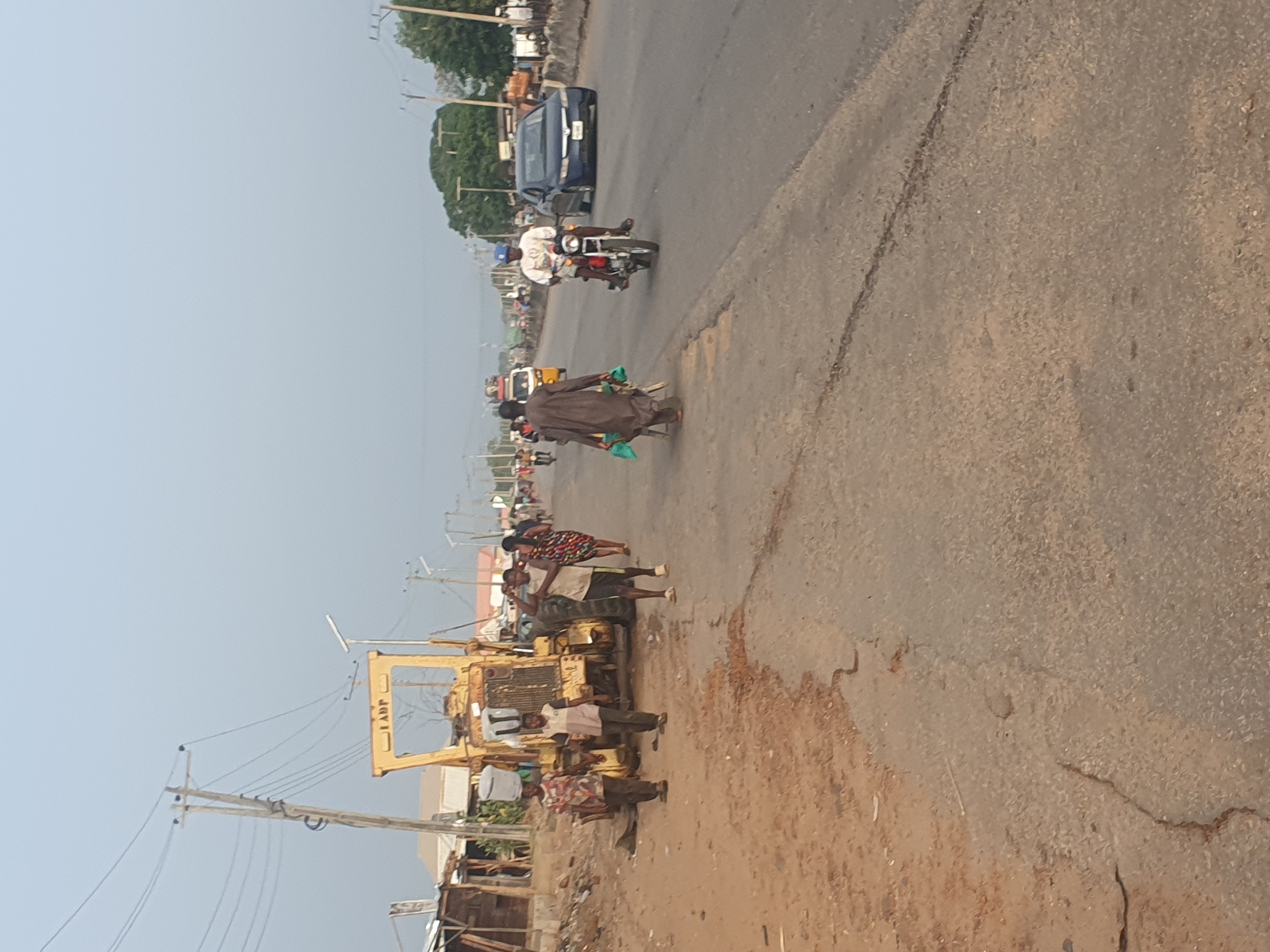 The image depicts a roadside scene in what appears to be Lokoja, Nigeria. A yellow road grader is parked on the left side of the road, with several people standing nearby, some carrying buckets on their heads. Another person in brown robes and carrying a green plastic bag is walking away. Traffic is flowing, with a car and a motorcycle with two people on it visible in the background. The road surface is cracked and worn. Buildings and power lines are also visible.