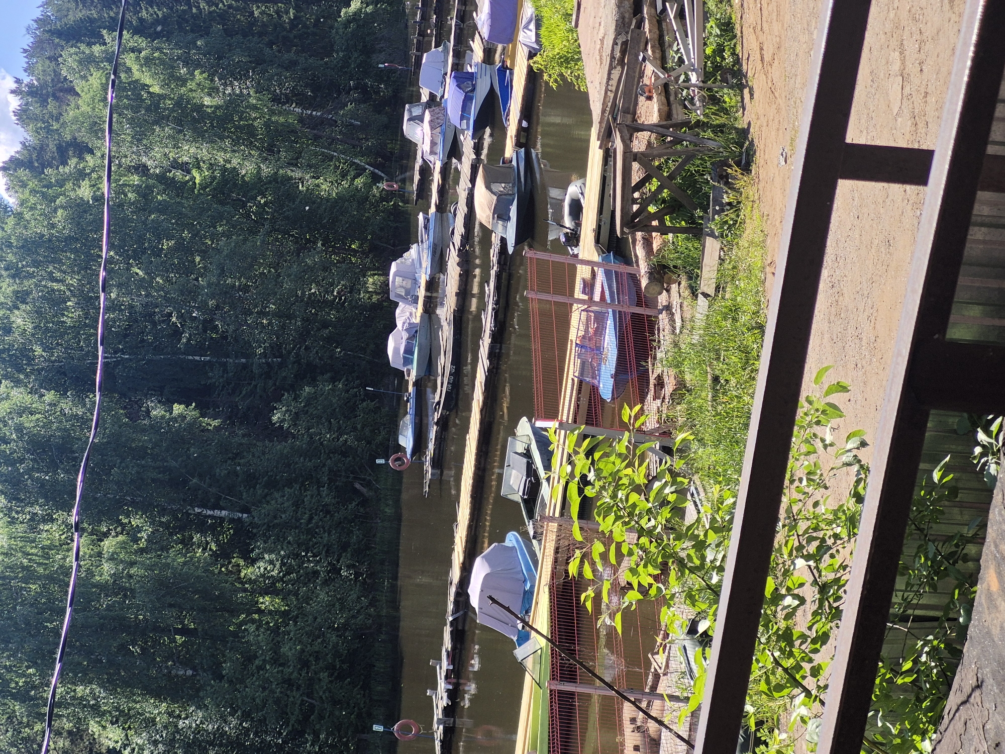 The image depicts a boat dock or marina situated along a waterway, possibly a river or canal. Multiple boats are moored along the dock, some covered with protective tarps. Trees densely line the opposite bank of the waterway, creating a lush, green backdrop. The foreground features a partially visible metal structure, likely part of a building or fence, obscuring some of the view. The scene appears to be set in the city of Chaikovsky, Russia, based on visual cues like the style of boats and architecture.