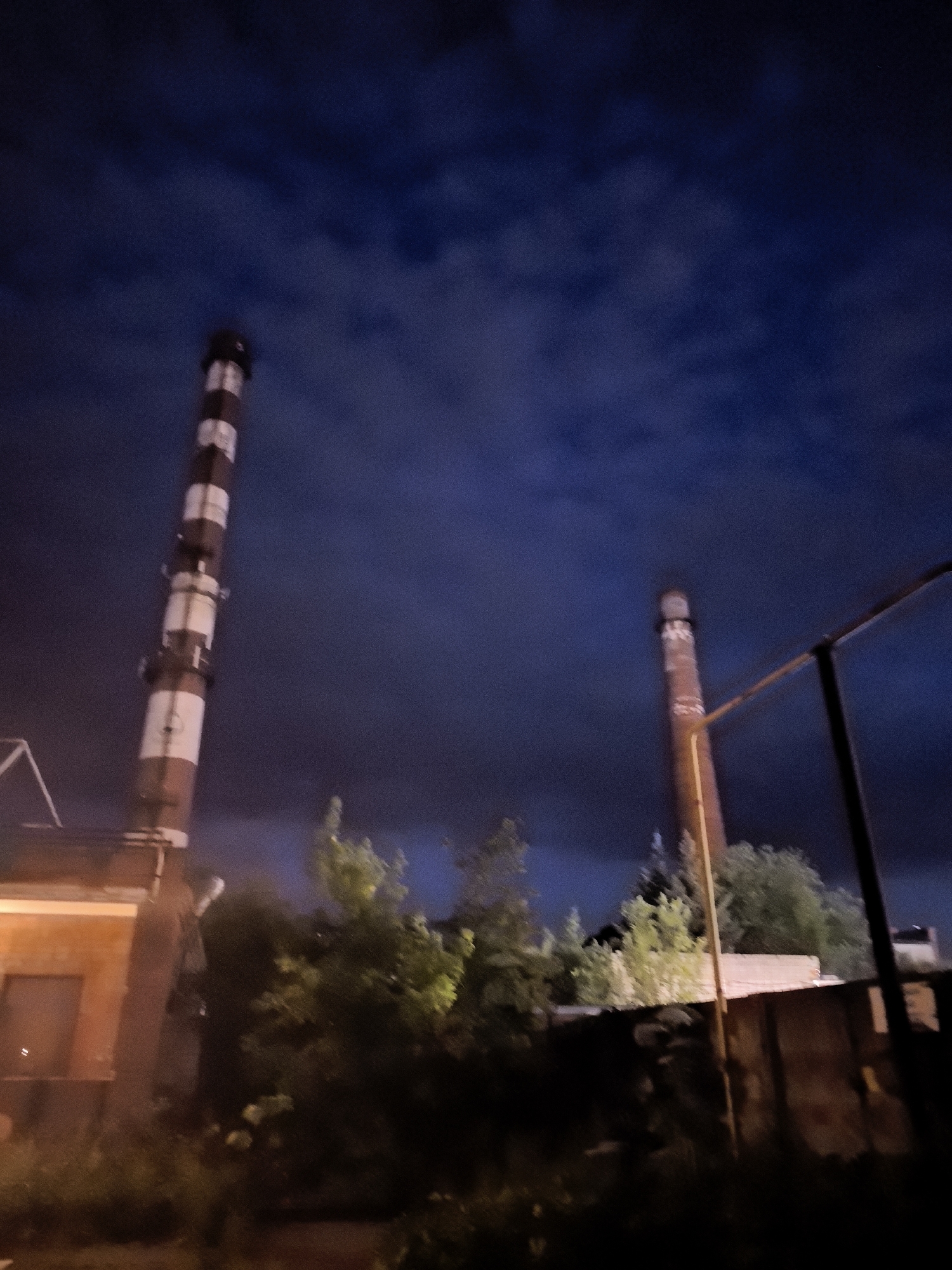 An outdoor, low-angle, night-time photograph captures a scene in Novo-Talitsy, Russia, featuring two industrial smokestacks and surrounding structures against a dark, cloudy sky. The larger smokestack on the left is tall and cylindrical, painted with alternating white and red horizontal bands, and appears to be made of brick. A portion of a brick building with a window is visible to the left of this smokestack, illuminated by a warm light source. In the mid-ground, dense green foliage and trees obscure the base of the smokestacks and other structures. A second, smaller smokestack, reddish-brown and cylindrical, is visible in the background to the right, partially obscured by trees. A rusty metal structure, possibly fencing or scaffolding, runs diagonally across the right side of the frame. The overall impression is one of an abandoned or disused industrial area.