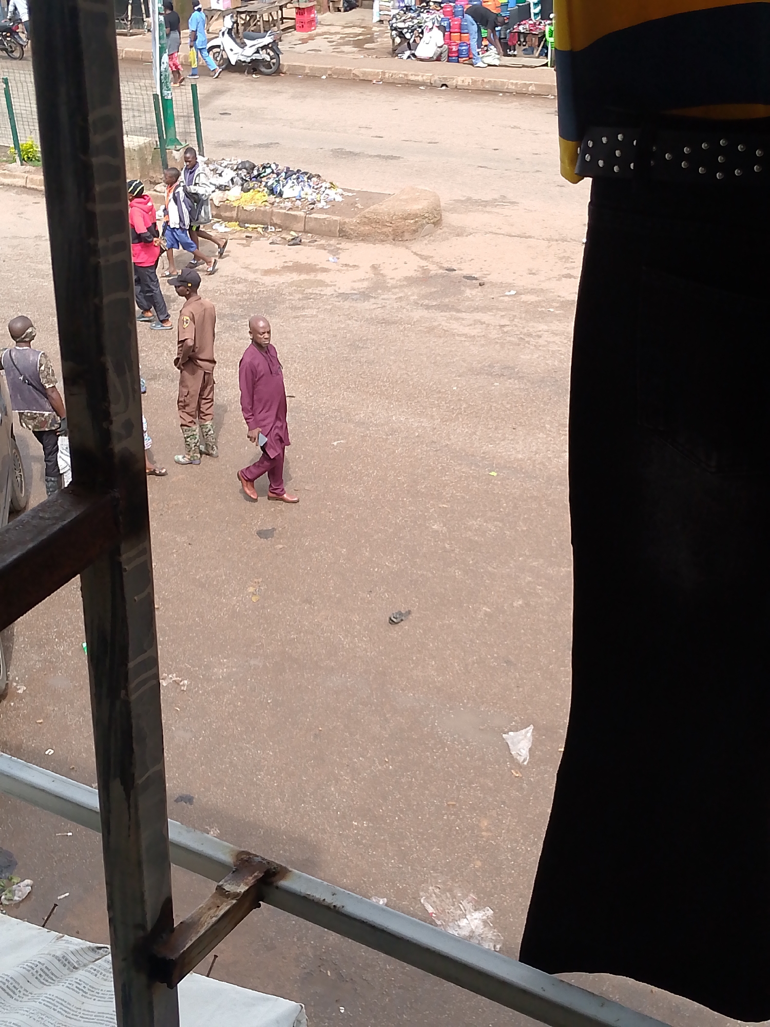 The image depicts a street scene in Bukuru, Nigeria, characterized by a dusty, reddish-brown ground and several individuals in casual and semi-formal attire. In the foreground, a black metal railing and a portion of a person wearing a black skirt and a yellow and blue striped top are visible. In the mid-ground, a man in a maroon tunic and pants walks away from the camera, holding a dark object in his right hand. To his left, another man in a tan uniform with camouflage boots stands with his hands behind his back. Further left, a person in a red hoodie and black pants walks away from the camera, accompanied by another individual wearing a blue and white jacket and blue shorts. Behind them, a pile of refuse is situated near a low wall. In the background, several motorbikes are parked, and in the far distance, a market stall with various goods is visible. The overall environment appears to be an outdoor marketplace or busy street area.