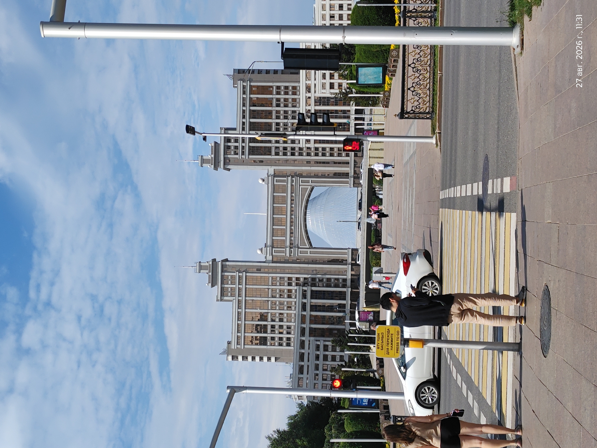 This image depicts a street scene in Astana, Kazakhstan, specifically in front of the Palace of Peace and Reconciliation. In the foreground, a pedestrian crossing is visible with yellow and white stripes. Two individuals are standing on the sidewalk near the crossing. One, a woman wearing a light brown tank top and black shorts, is looking away from the camera. The other, a man in a black hooded sweatshirt and beige pants, is looking down at his phone. A white car is stopped at the crossing, and a traffic light displays a red signal for pedestrians. A yellow pedestrian signal button with text in Kazakh and Russian is attached to a pole. Behind the pedestrian crossing, a large, multi-story building with a distinctive arched entrance and a conical-shaped structure inside can be seen. This is the Palace of Peace and Reconciliation. The sky is blue with scattered white clouds. The overall scene is that of a sunny day in a city environment.