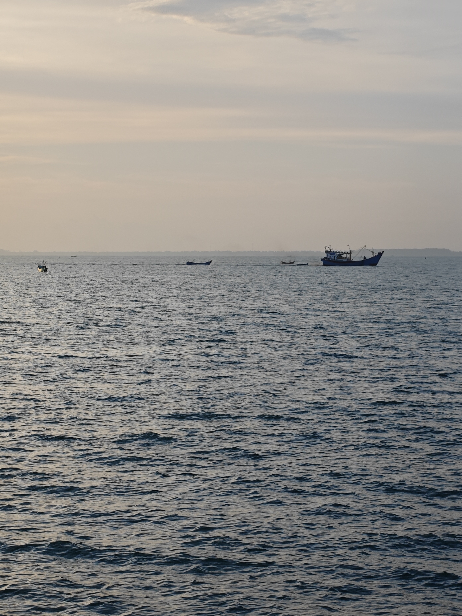 The image depicts a seascape off the coast of Lhokseumawe, Indonesia. The scene is dominated by the sea, with gentle waves covering the surface. Several boats are visible on the horizon, including a larger fishing vessel with visible rigging and a few smaller boats scattered across the water. The sky is overcast with soft, muted colors suggesting either early morning or late afternoon. There are no visible people or specific activities depicted, focusing solely on the boats and the marine environment.