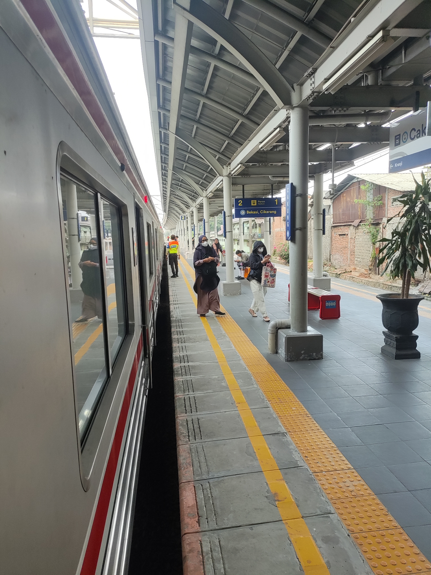 The image captures a train station platform in Bekasi, Indonesia. A train is stopped alongside the platform, with its doors open. Several people are visible on the platform. One person in a high-visibility vest stands near the train, possibly a station attendant. Other individuals, some wearing masks, are either walking or standing, appearing to wait for or disembark from the train. The platform is paved with gray tiles and features a tactile warning strip and a yellow line marking the edge. Overhead, a gray metal roof supported by pillars shelters the platform. Signage is visible, including one indicating "Bekasi, Cikarang" and another pointing towards "Kranji". A potted plant is situated on the platform, and red benches are also present. The background shows some brick buildings and greenery.