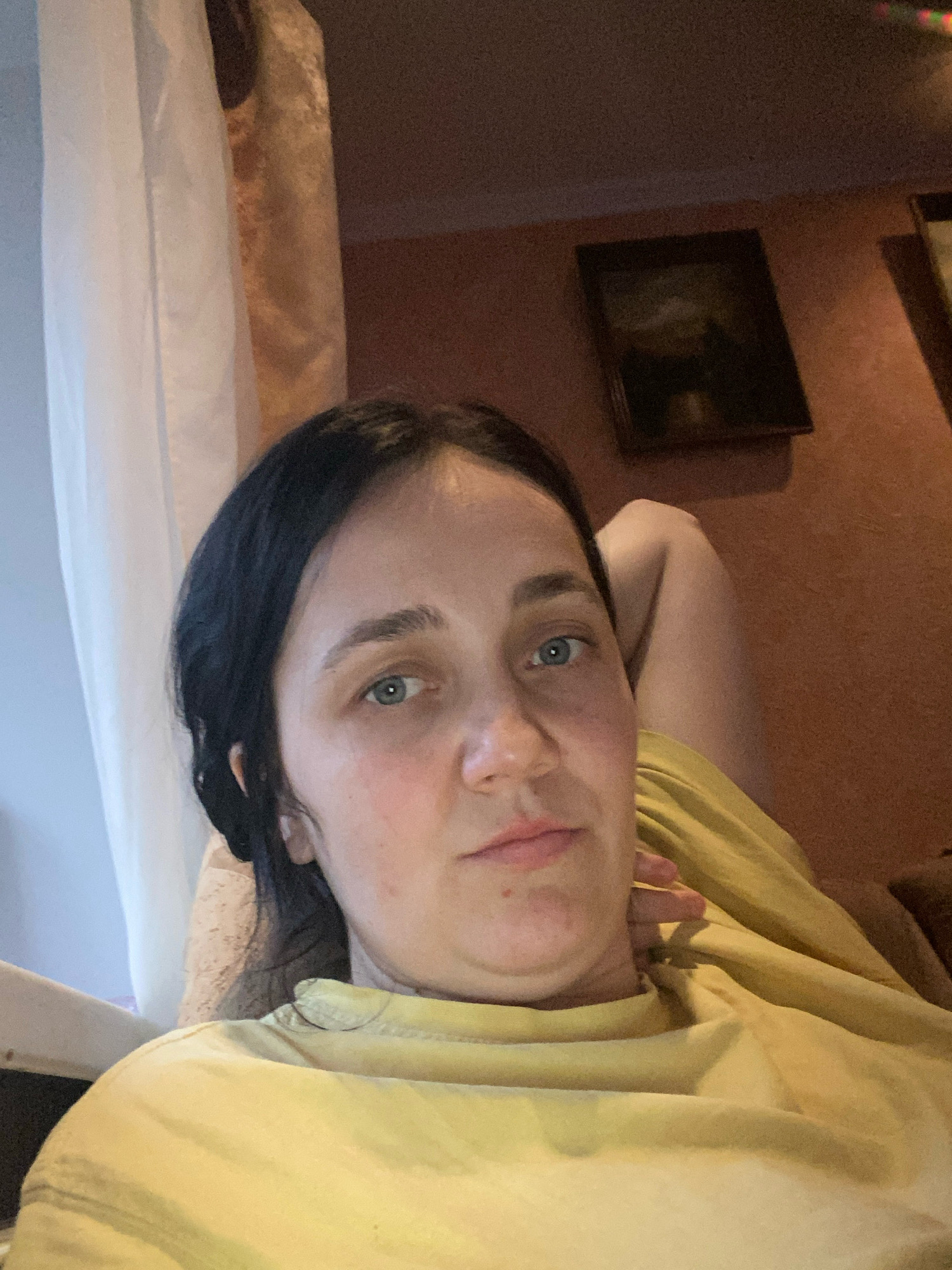 A woman with dark hair and blue eyes takes a selfie. She is wearing a yellow shirt and has her hair pulled back. She is lying down, with her arm bent and resting on her head. The background is a room with a curtain and a painting on the wall. The lighting is dim and warm, suggesting it is evening or indoors.
