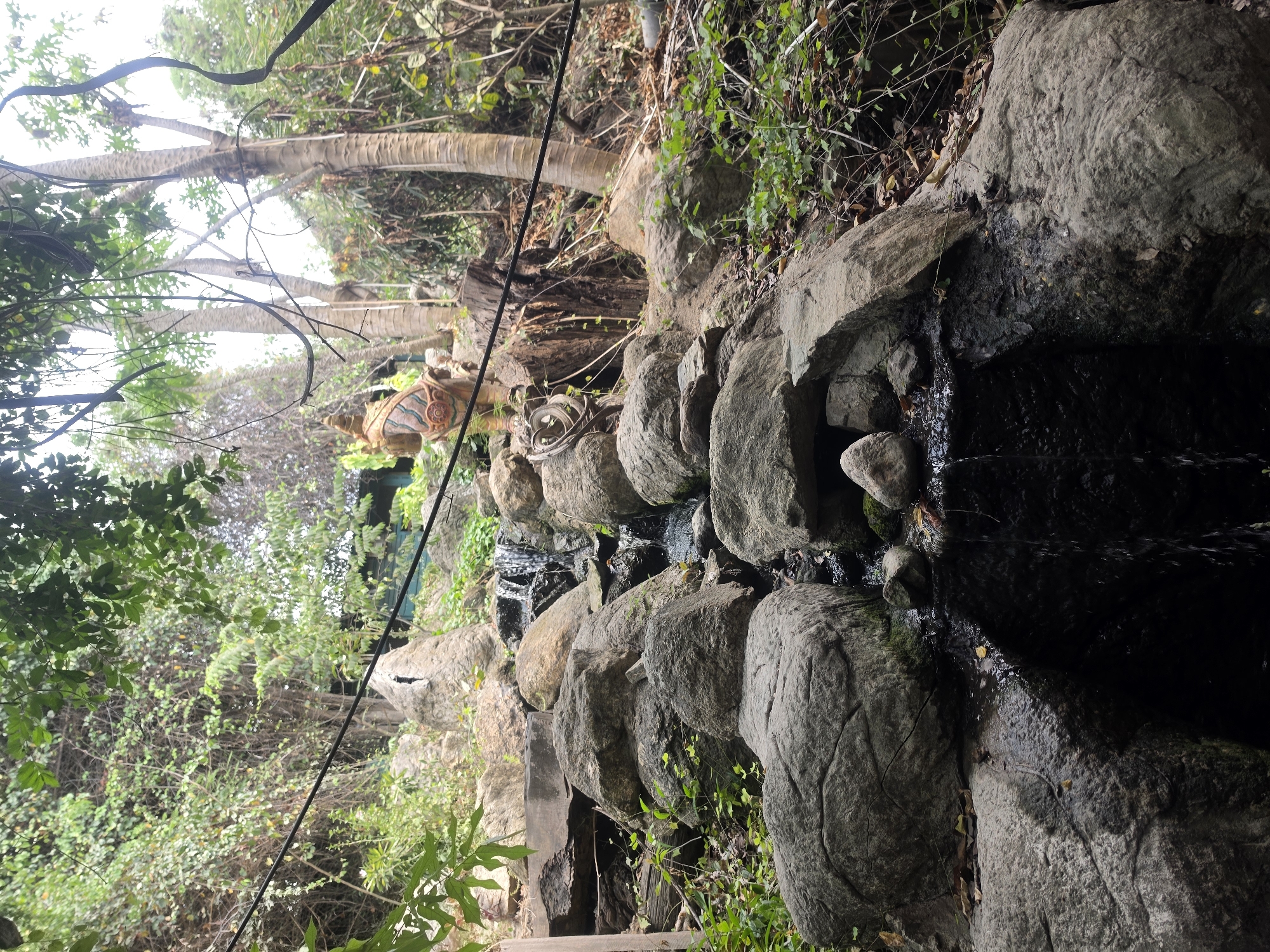 The image depicts a man-made waterfall constructed with large stones and boulders in Pattaya City, Thailand. The water flows down the rocks, creating a small stream. In the background, there are trees and vegetation. A decorative statue, possibly a horse or mythological creature, is situated on the embankment behind the waterfall. A power line is visible overhead, cutting diagonally across the frame.